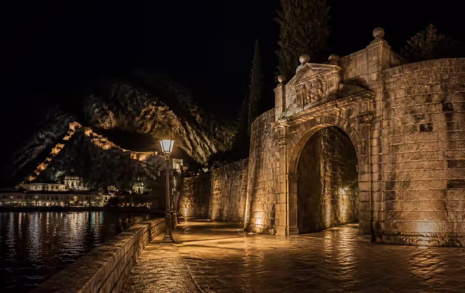 An old stone fortress gate and riverside promenade lit warm gold at night in a southern Balkan city