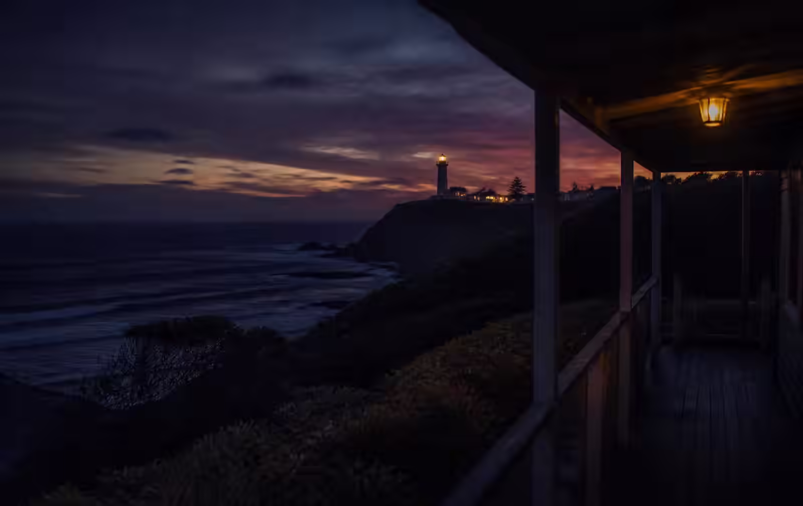 A dark subtropical coastline at dusk with a distant lighthouse headland seen from a lamplit timber verandah