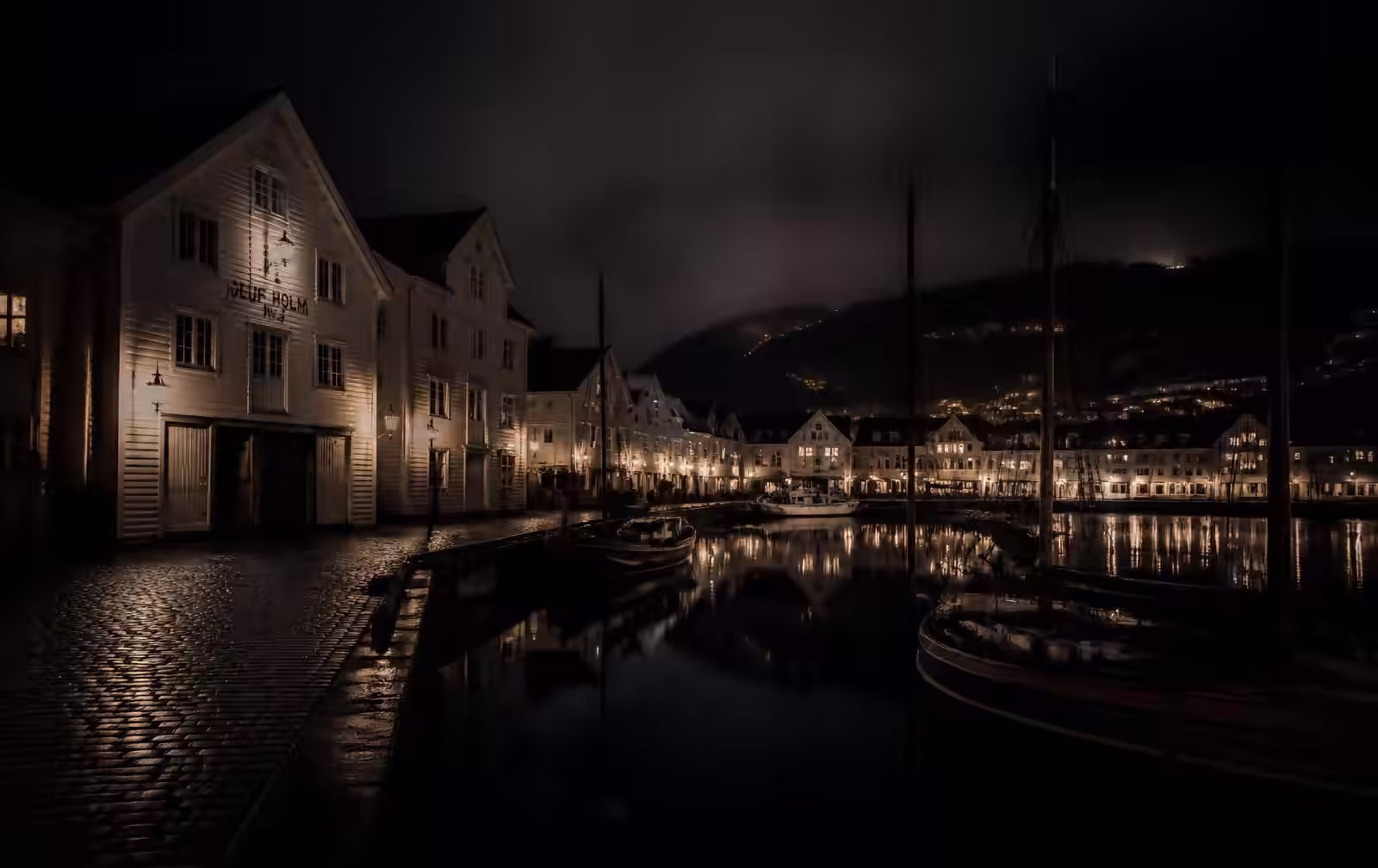 A Norwegian coastal city harbour at night, white wooden warehouse buildings lit by warm lamps, moored boats in silhouette on black water