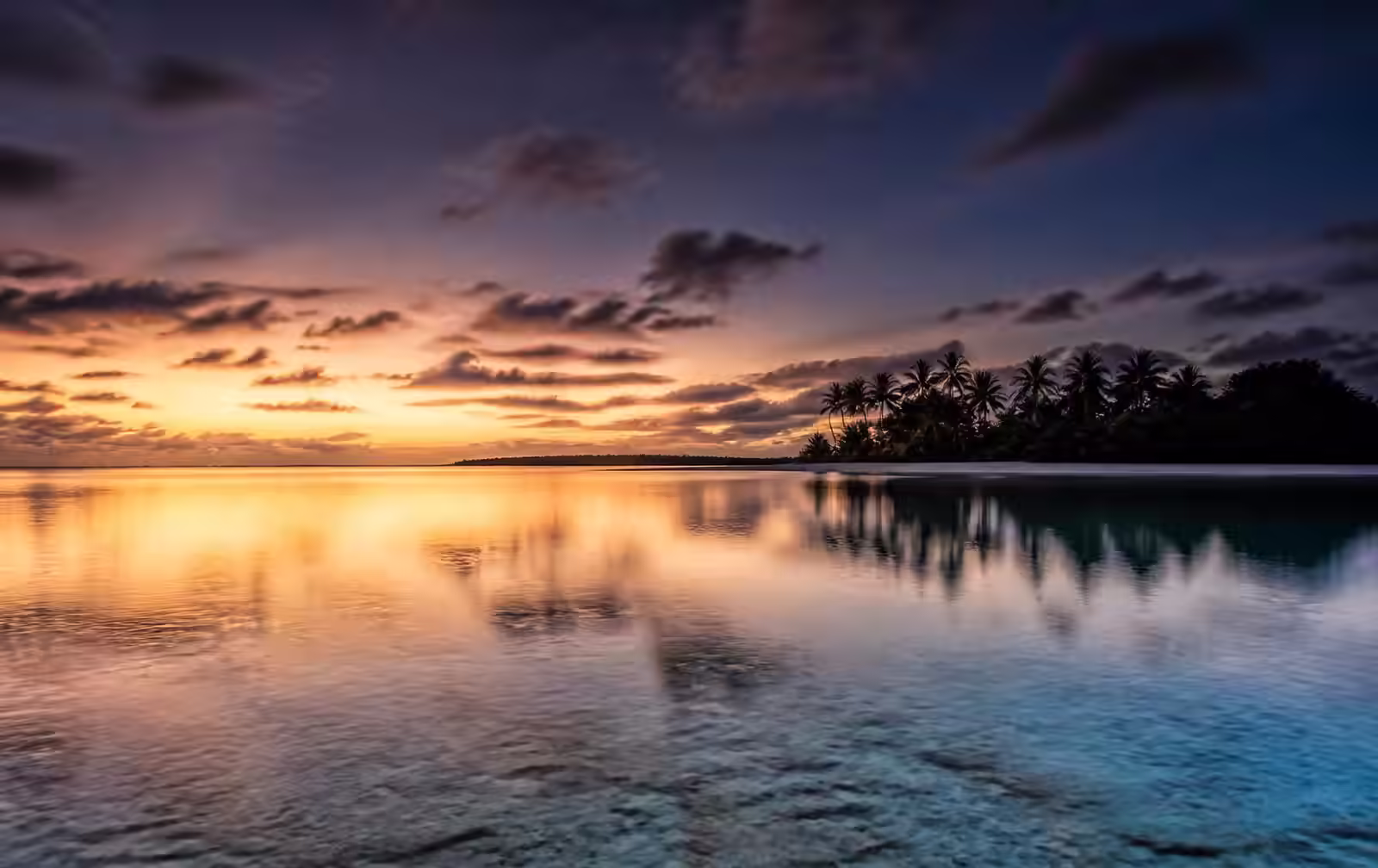 A wide flat lagoon at dusk reflecting pale gold light with a low palm island on the horizon