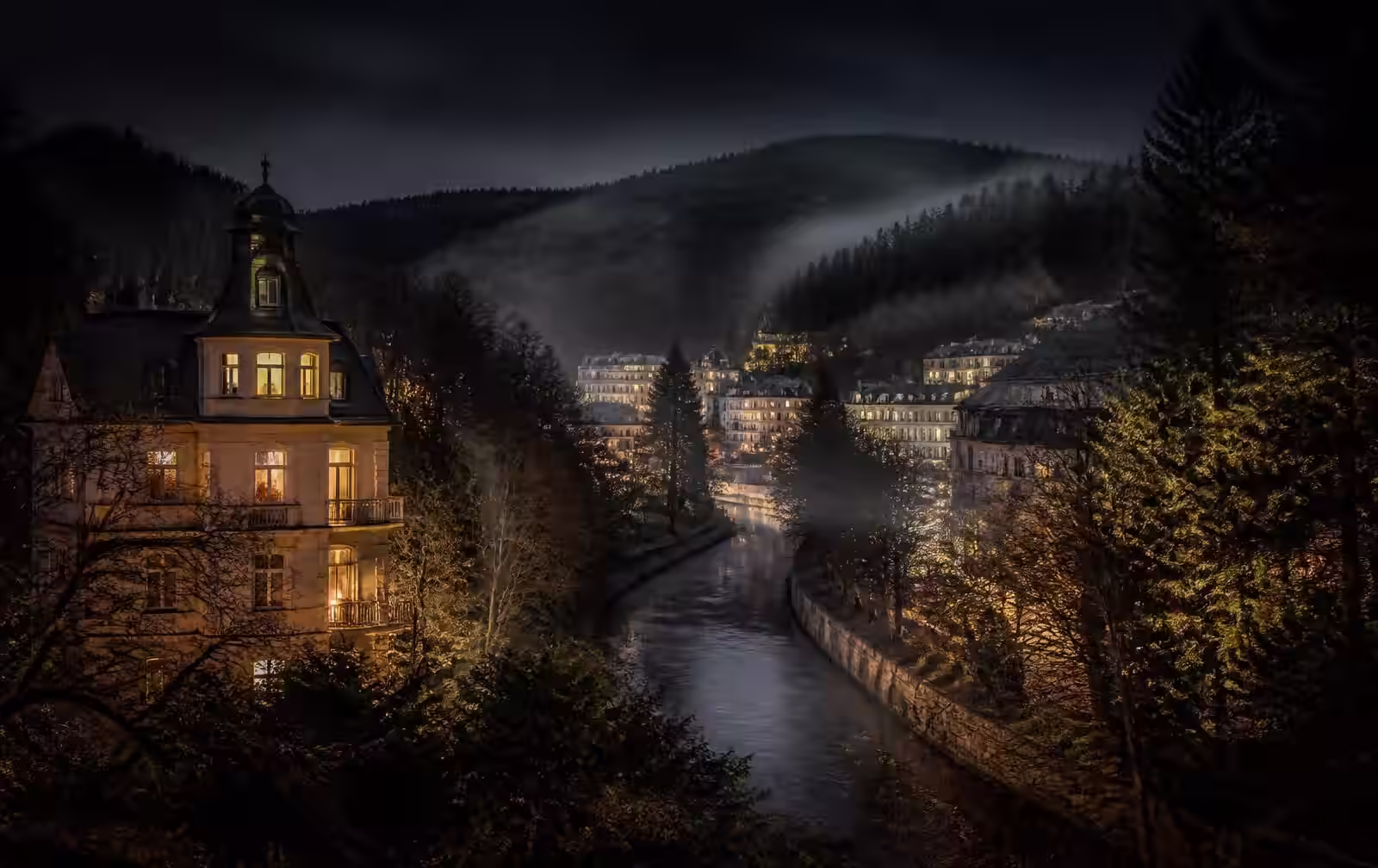 A small wooded spa town in a misty valley at night with old villas lit warm gold beside a stream