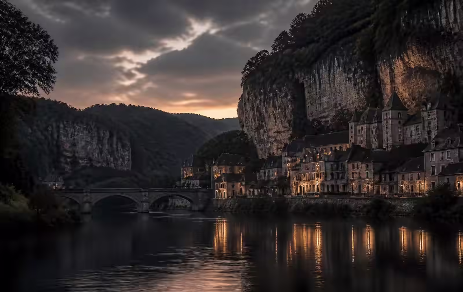 Broad Meuse river at dusk between wooded limestone cliffs with an old stone town of lit slate-roofed houses along the water