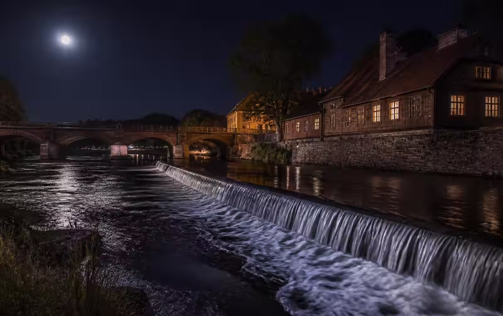 Kuldīga at night — the wide Venta waterfall glimmering under moonlight, an old brick bridge and warm-lit timber houses