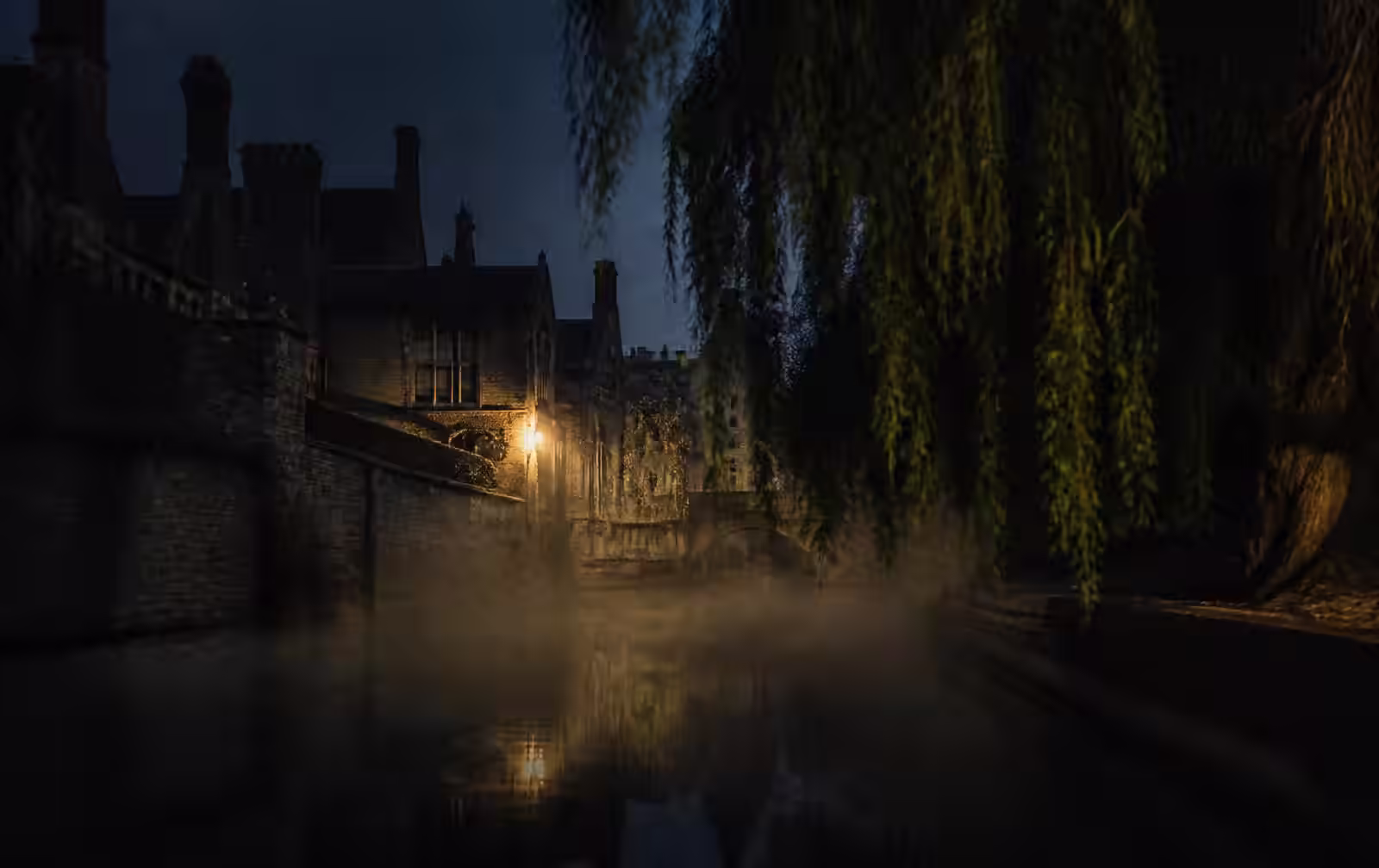 Misty river at night beside old English college stone walls with willows and one warm light