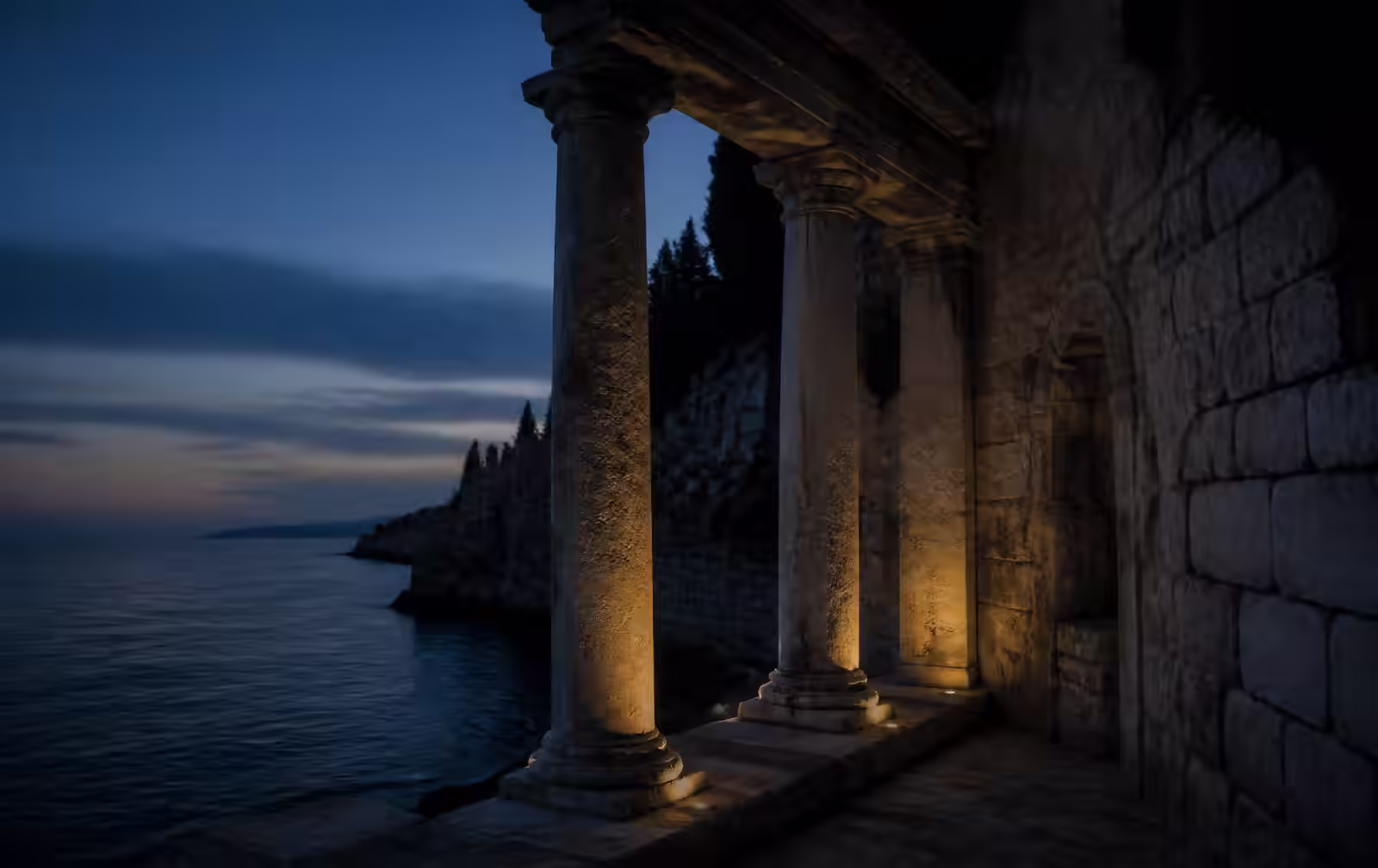 Ancient stone columns and a ruined coastal wall beside a dark Mediterranean sea at blue hour