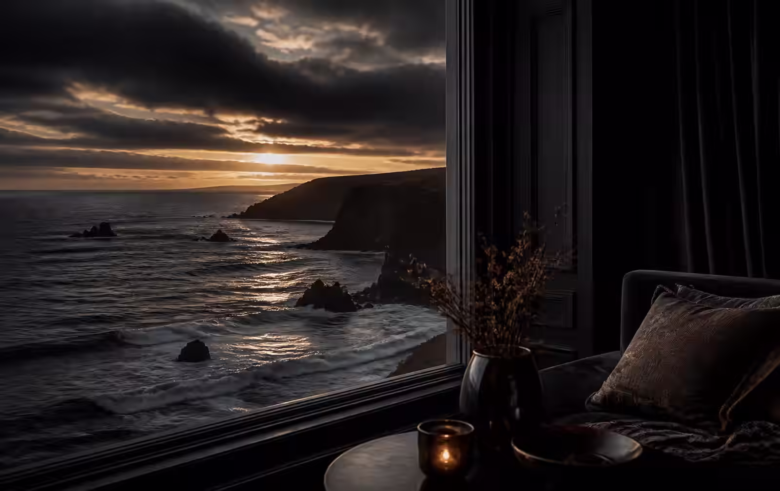 A dark Atlantic coastline at dusk seen from a refined interior with candlelight and deep shadow