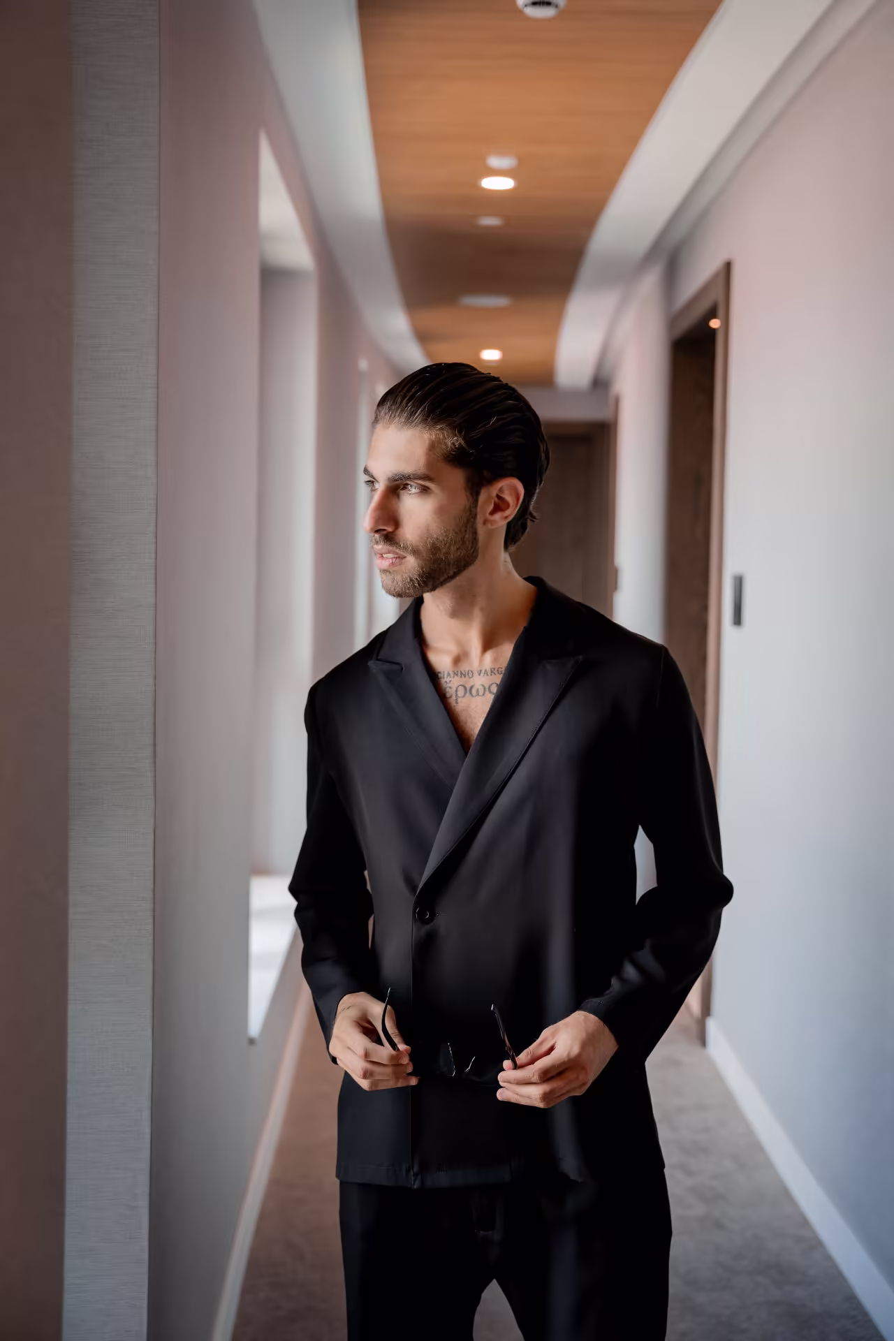 Lucianno Vargas in a black blazer in a softly lit hotel corridor, sunglasses folded in his hands