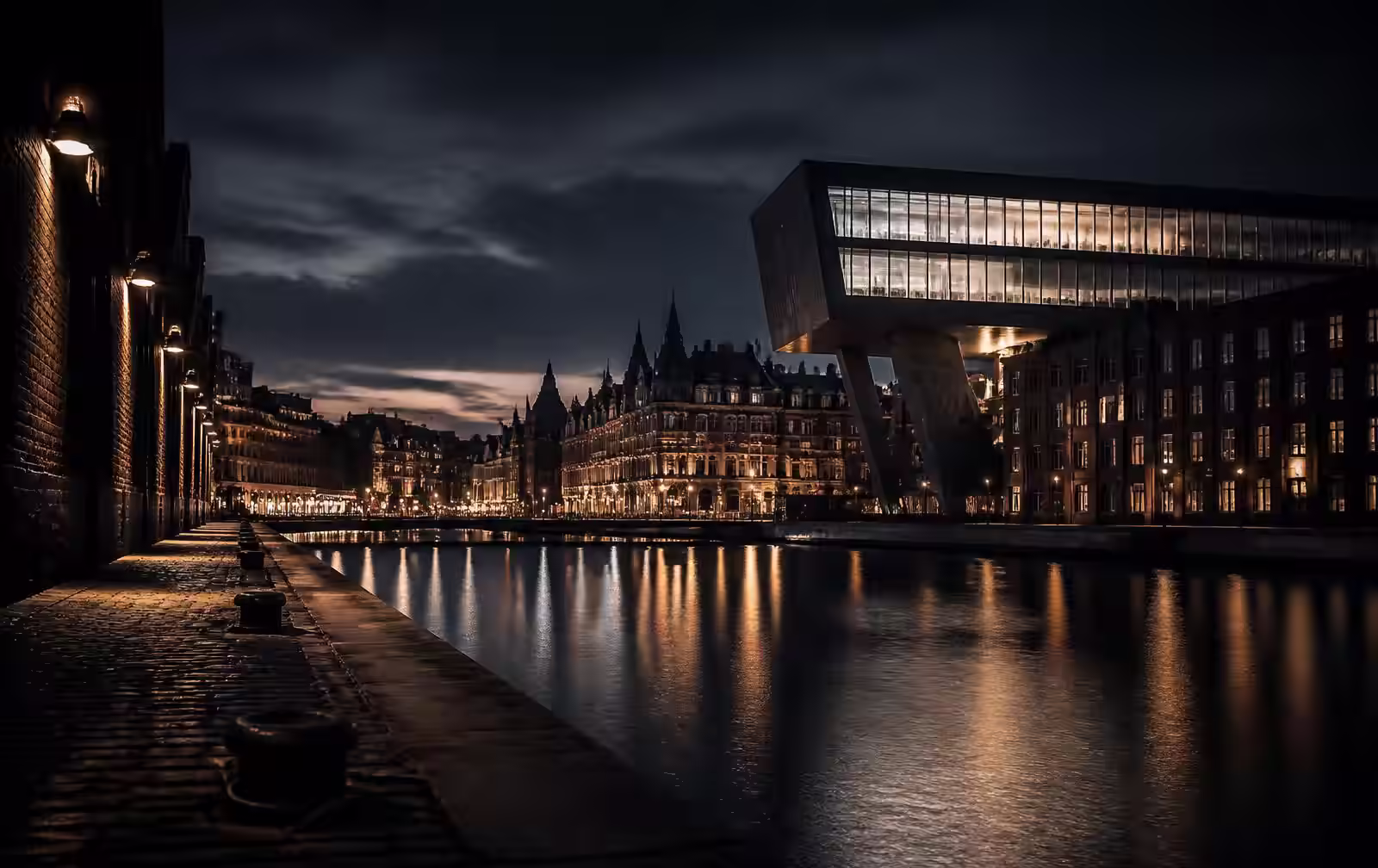 Antwerp river quay at night with historic warehouse façades, bold modern architecture and lights reflecting on dark water