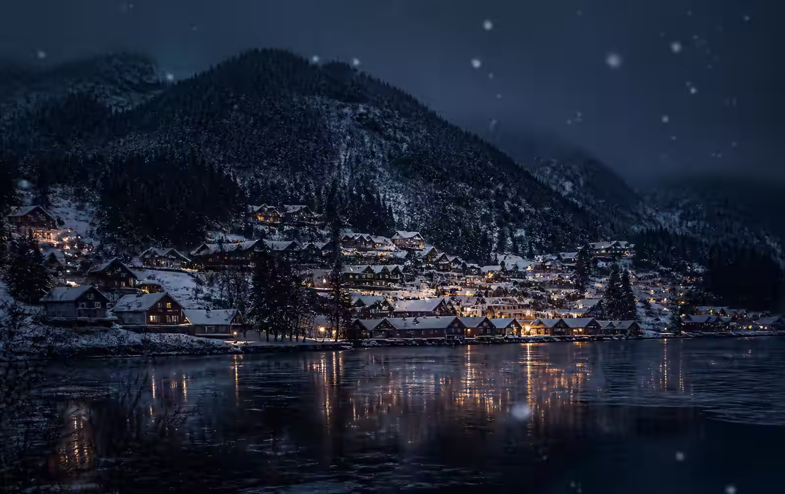 An inland Norwegian mountain town at night in falling snow, wooden buildings with warm lit windows above a dark lake, forested hills behind