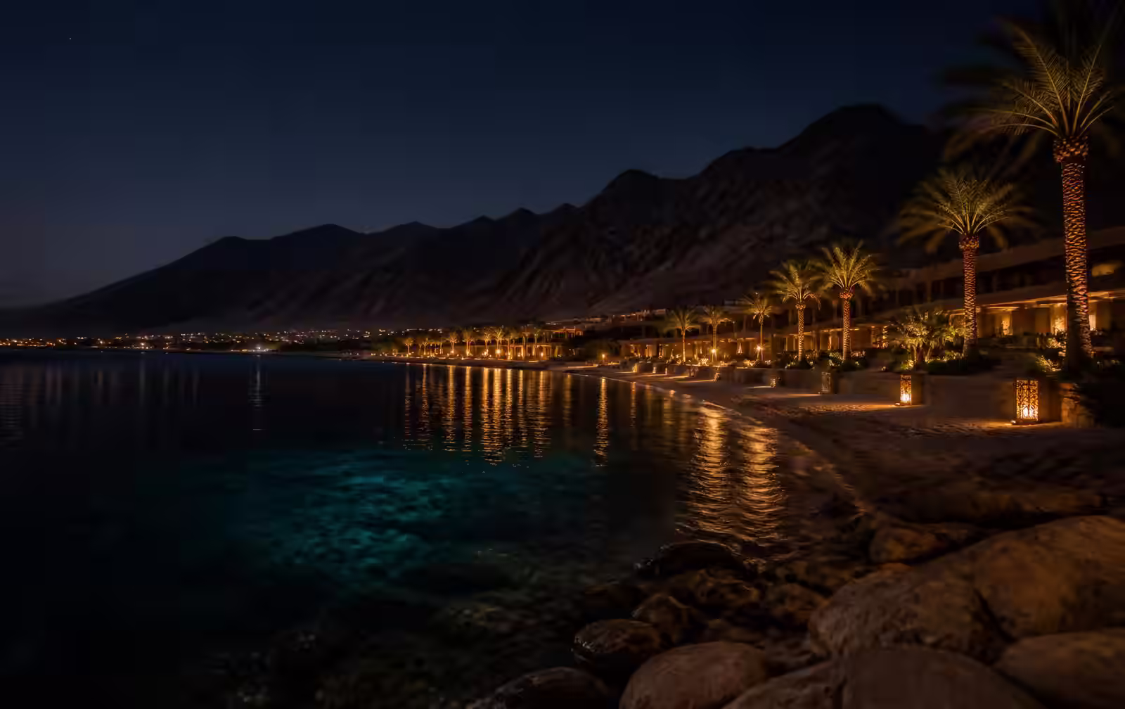 Red Sea bay at night with dark arid mountains behind a lit resort shoreline and still water