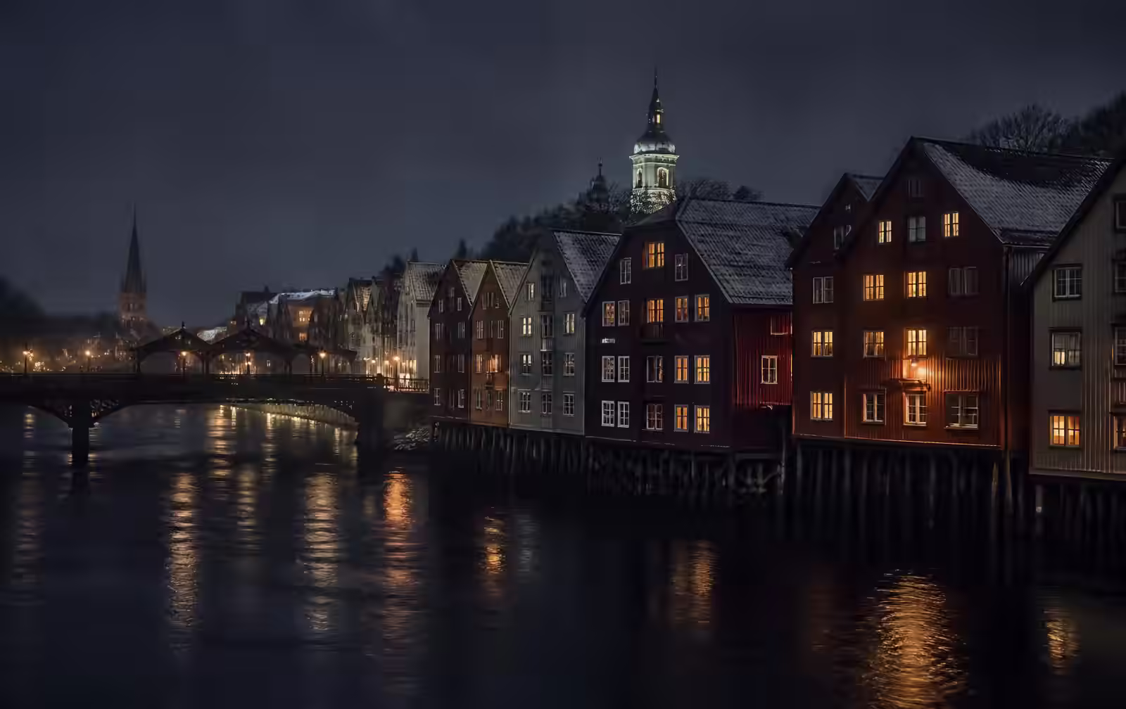 Colourful old wooden wharf buildings on stilts above a dark Norwegian river at night, warm window light reflected, an old bridge in silhouette