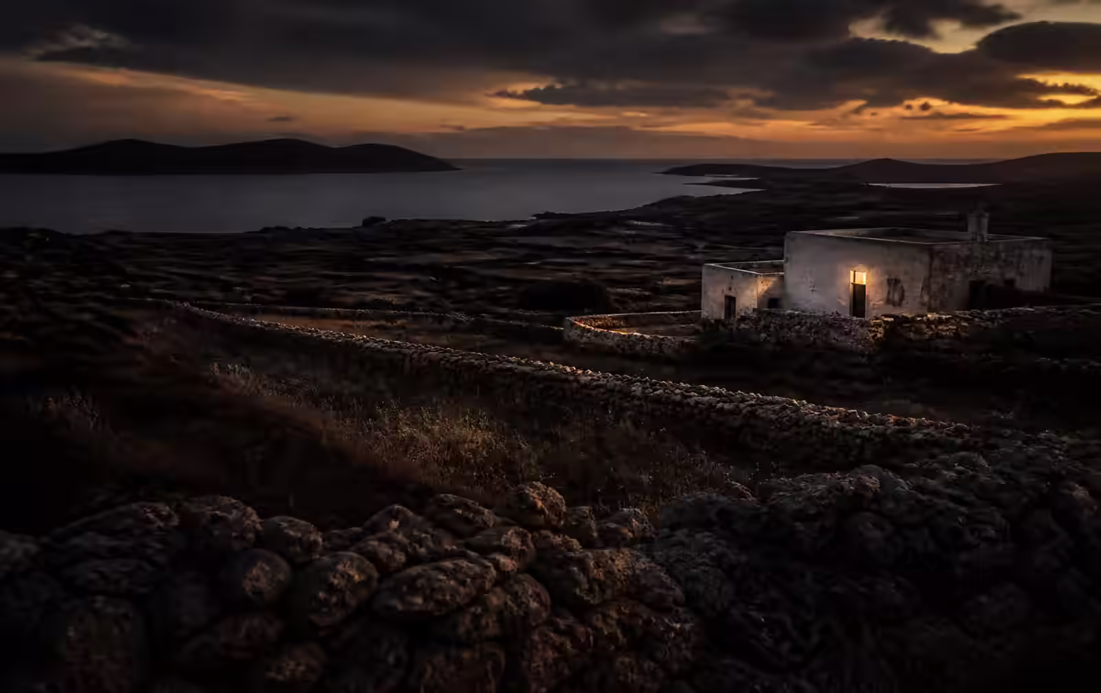 A quiet island landscape at dusk with dry stone walls, low countryside and a limestone farmhouse with one lit window near the sea