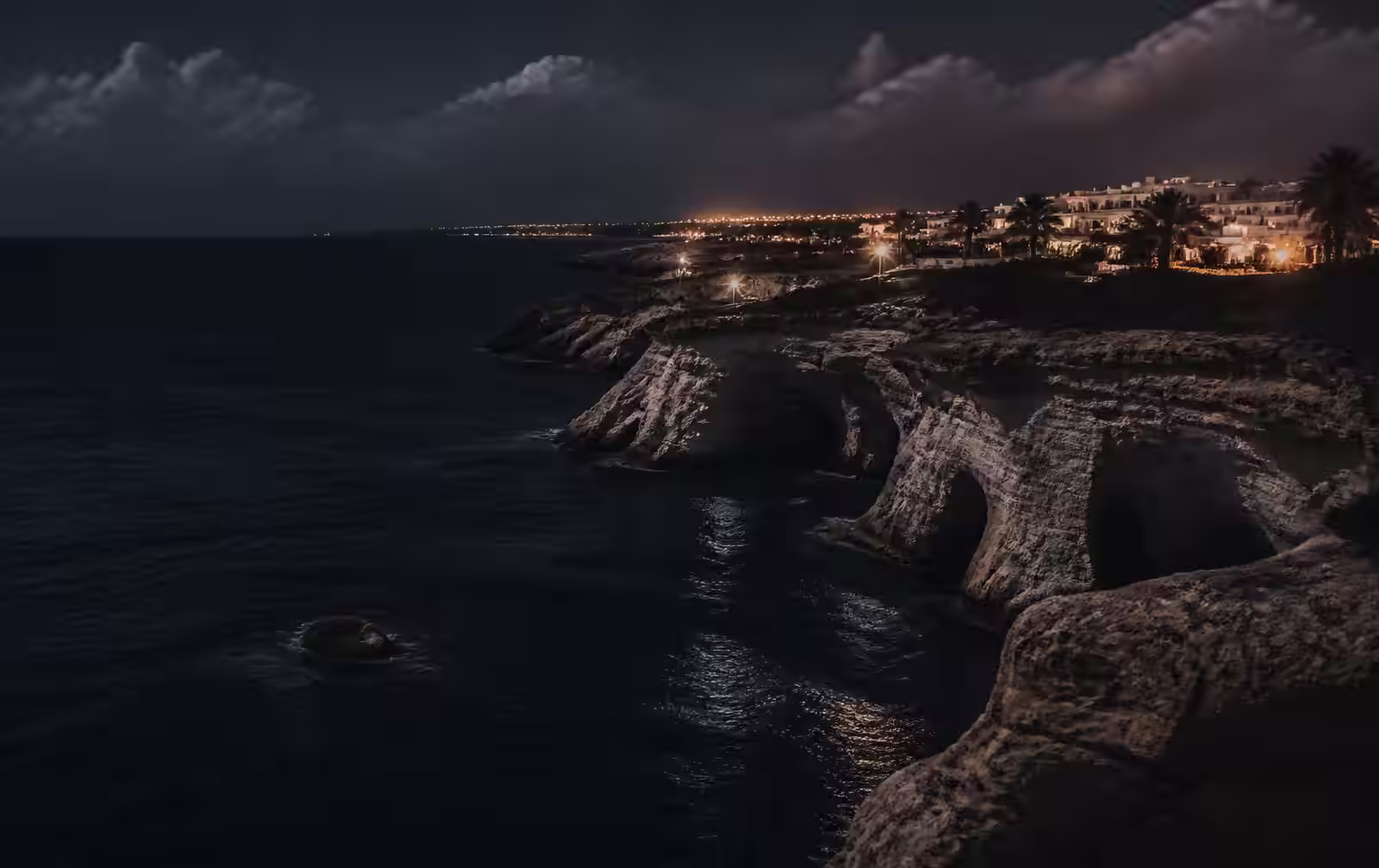 South-east Cyprus limestone sea caves at night with dark water and distant warm resort lights