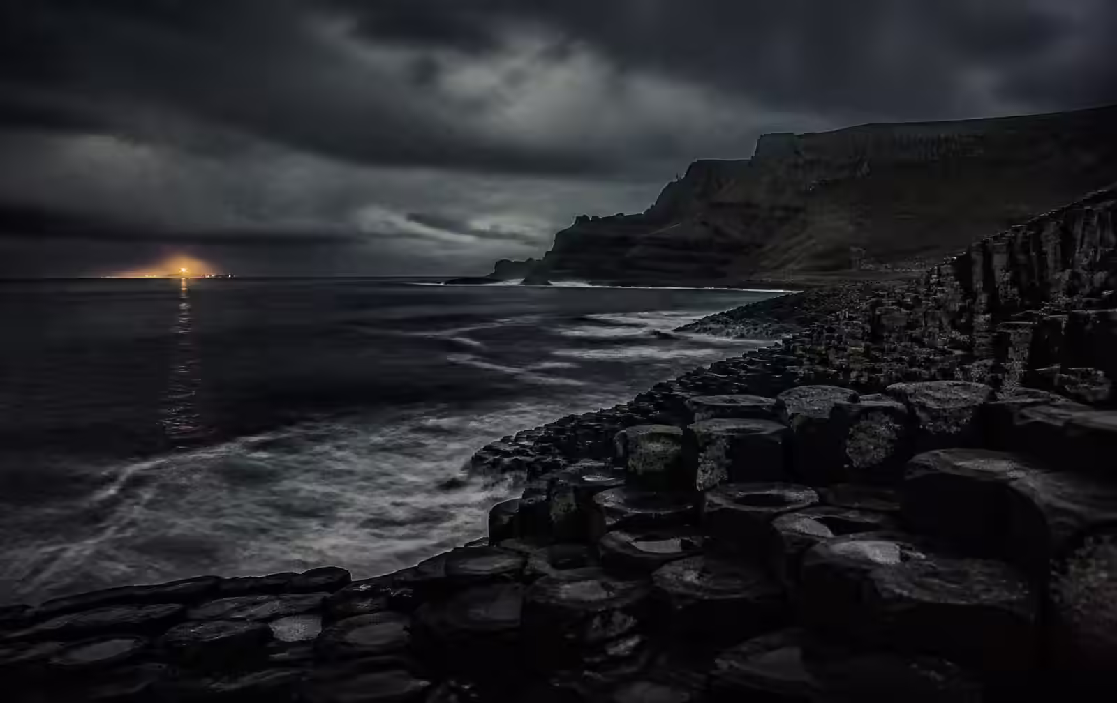 Dark basalt coastline of Northern Ireland at night under low cloud with a distant harbour light