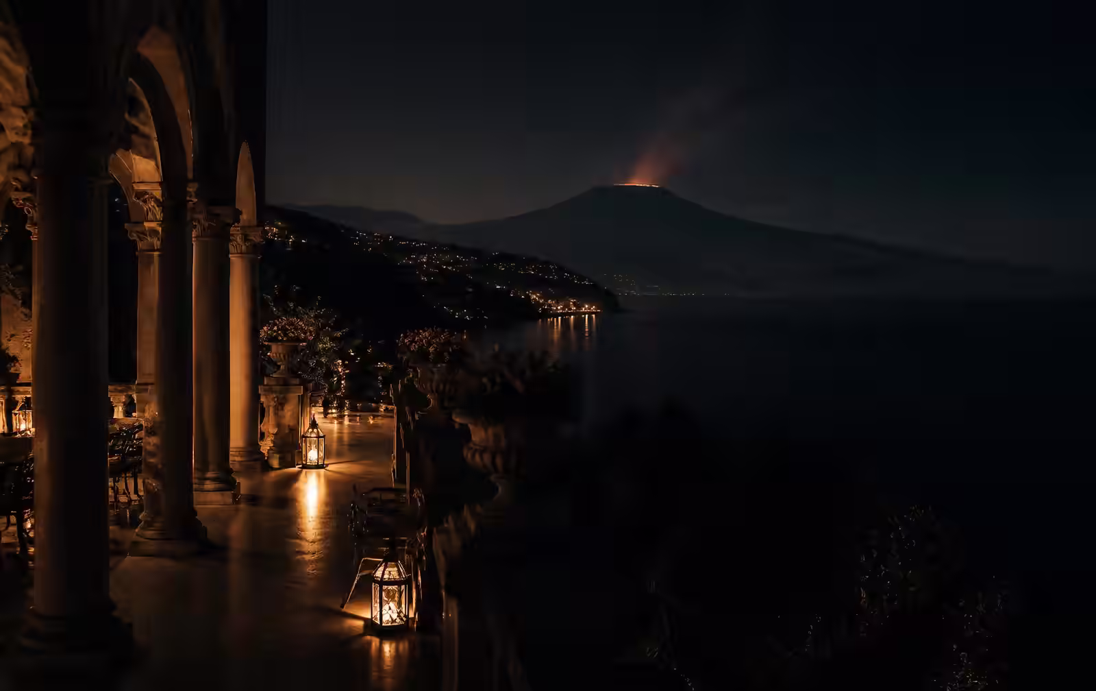 A columned Taormina terrace at night with lanterns, the dark Ionian sea below and Etna's silhouette beyond