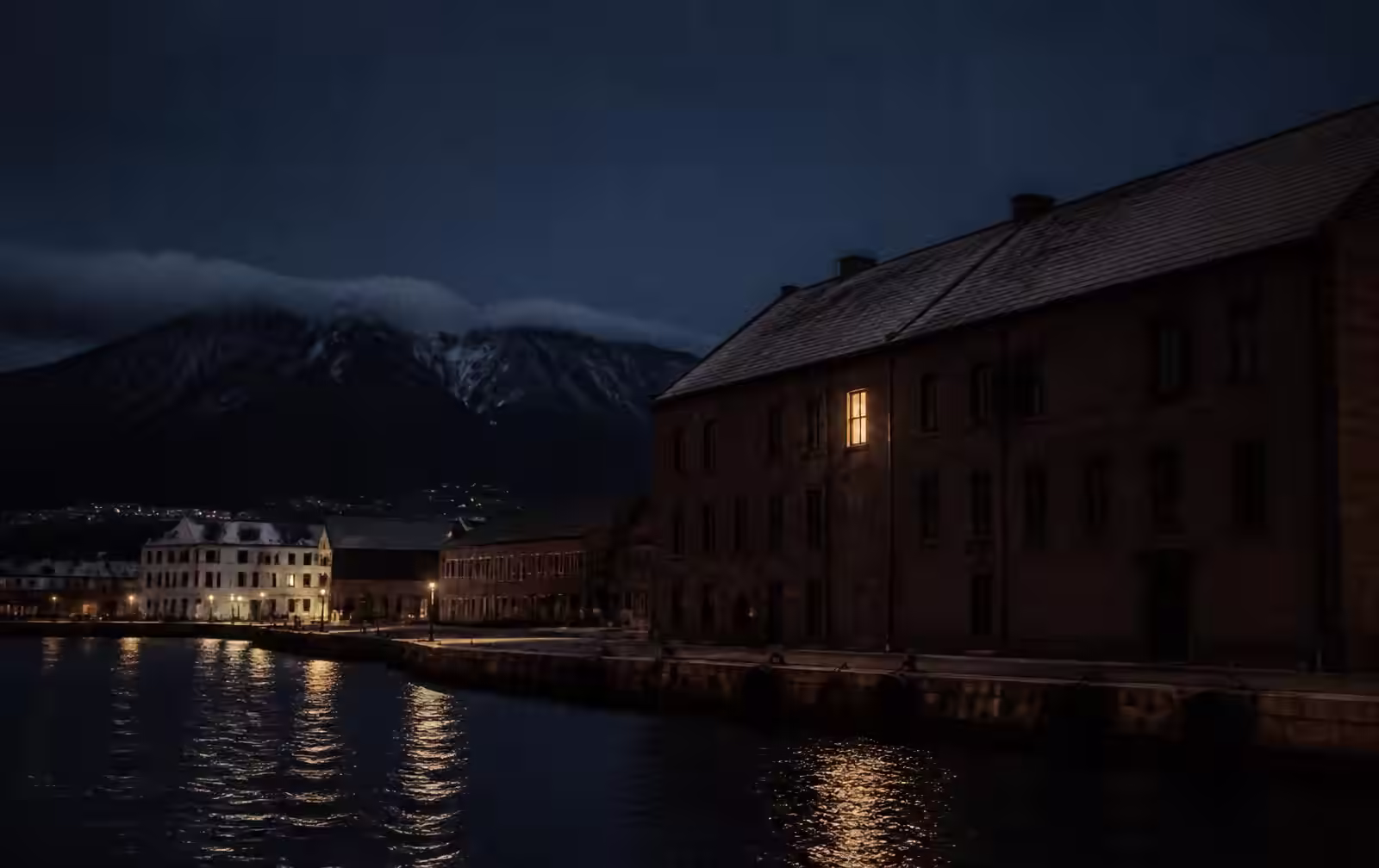 Hobart's waterfront at night with dark water, sandstone warehouses, a mountain silhouette and one warm lit window