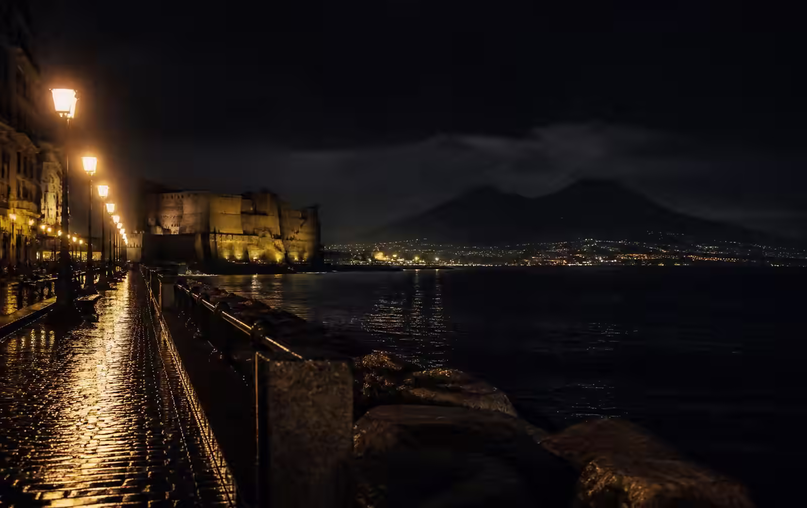 The Naples seafront at night with lamplit promenade, a floodlit seaside castle and Vesuvius across the dark bay