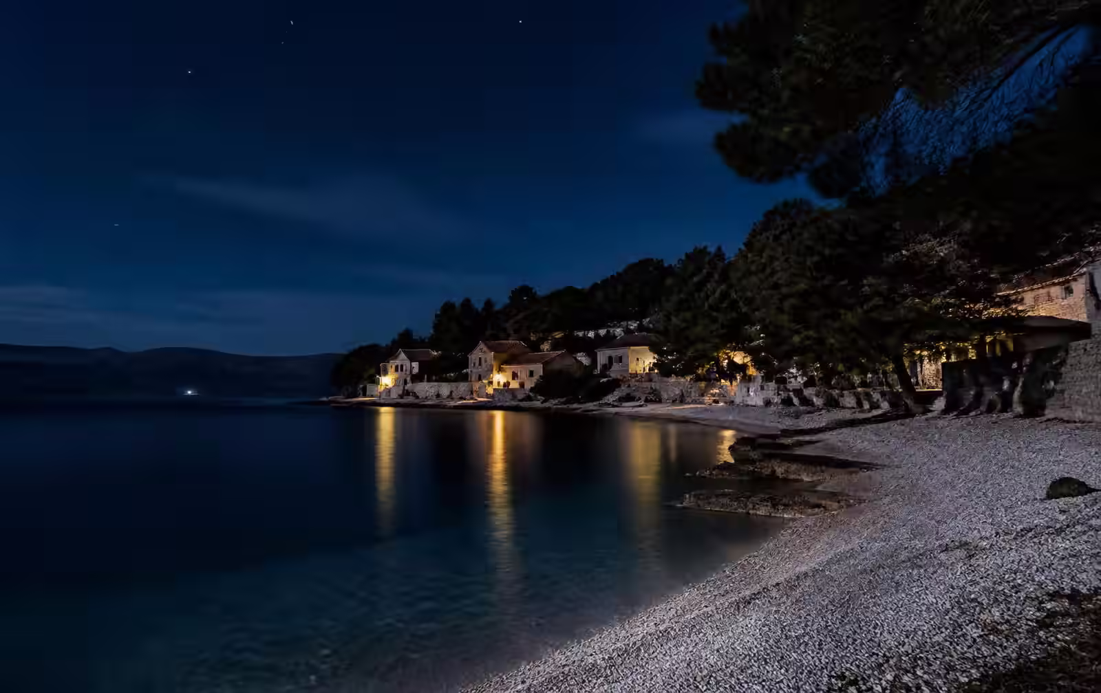 A quiet Croatian island cove at night — pale pebble shore, warm village lights, pine silhouettes under a starlit sky