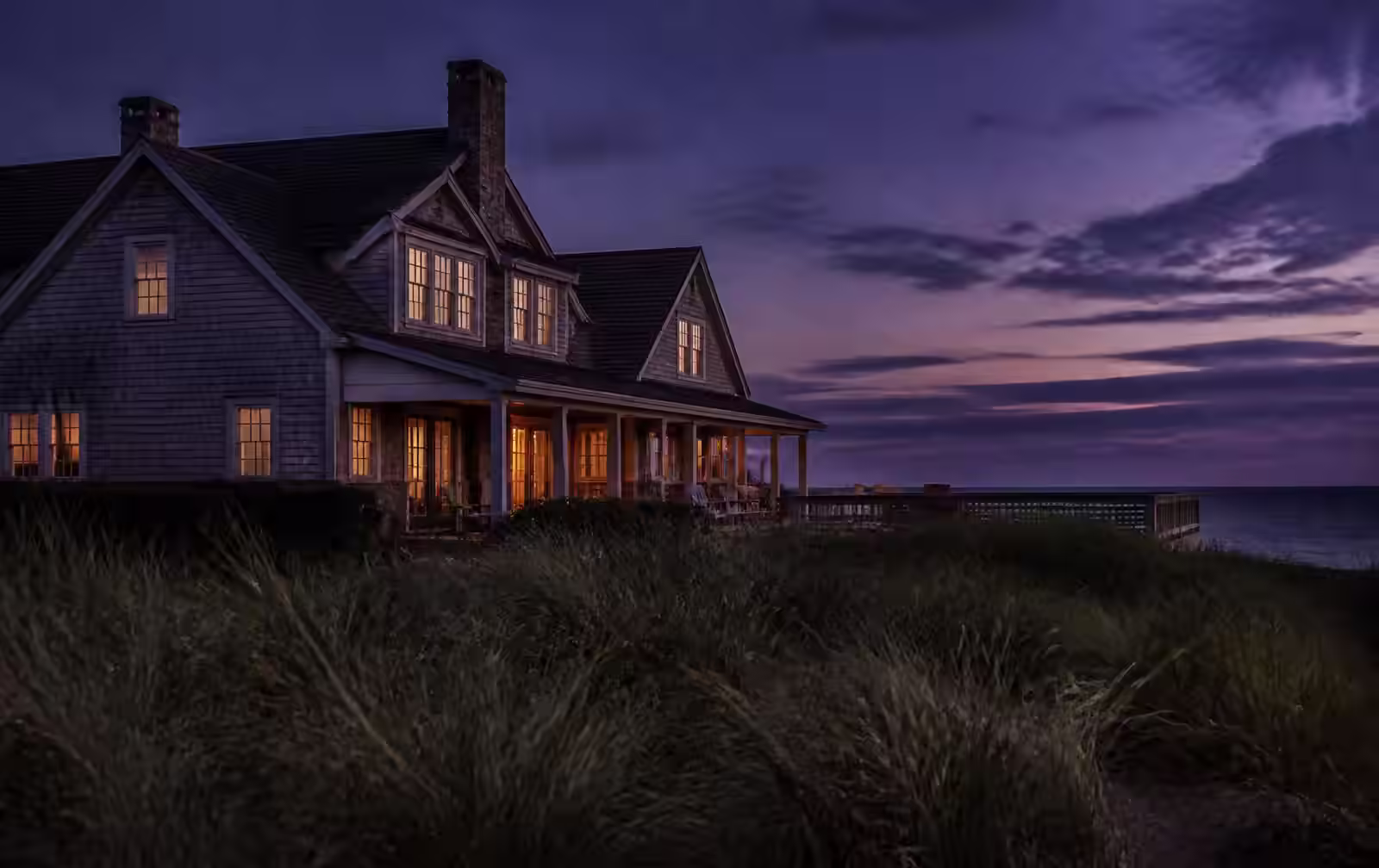 Shingled Hamptons house with warmly lit windows behind dune grass at summer dusk