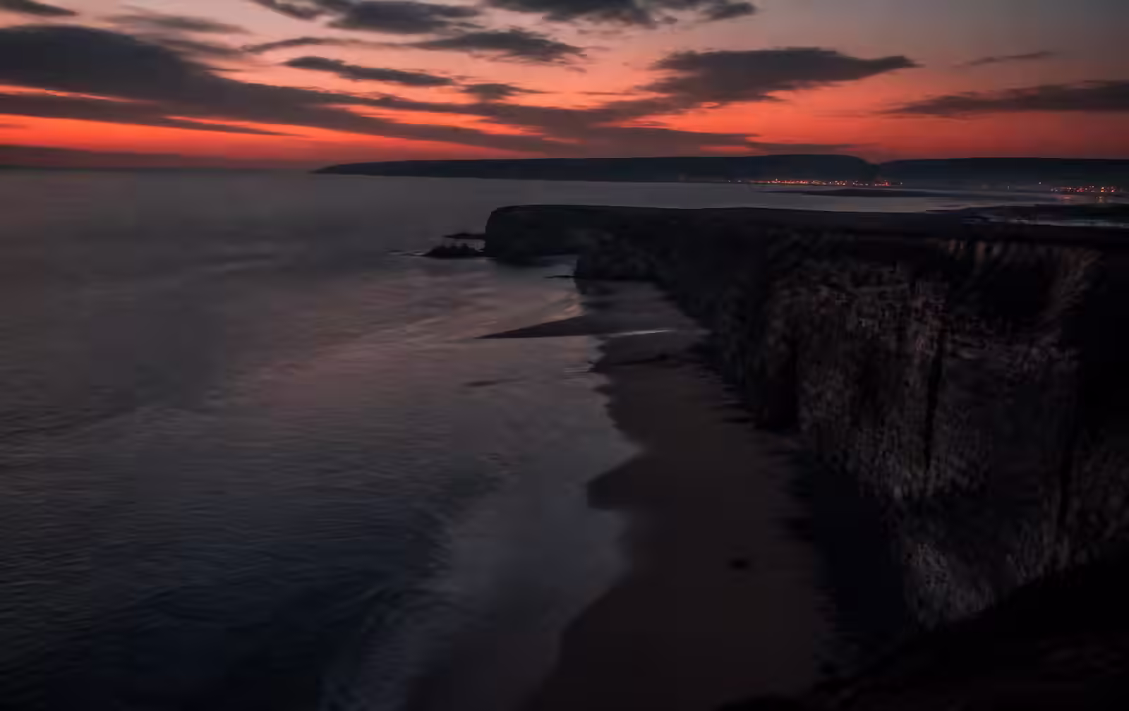 Welsh coastal cliffs at dusk above an empty beach with distant town lights along the bay