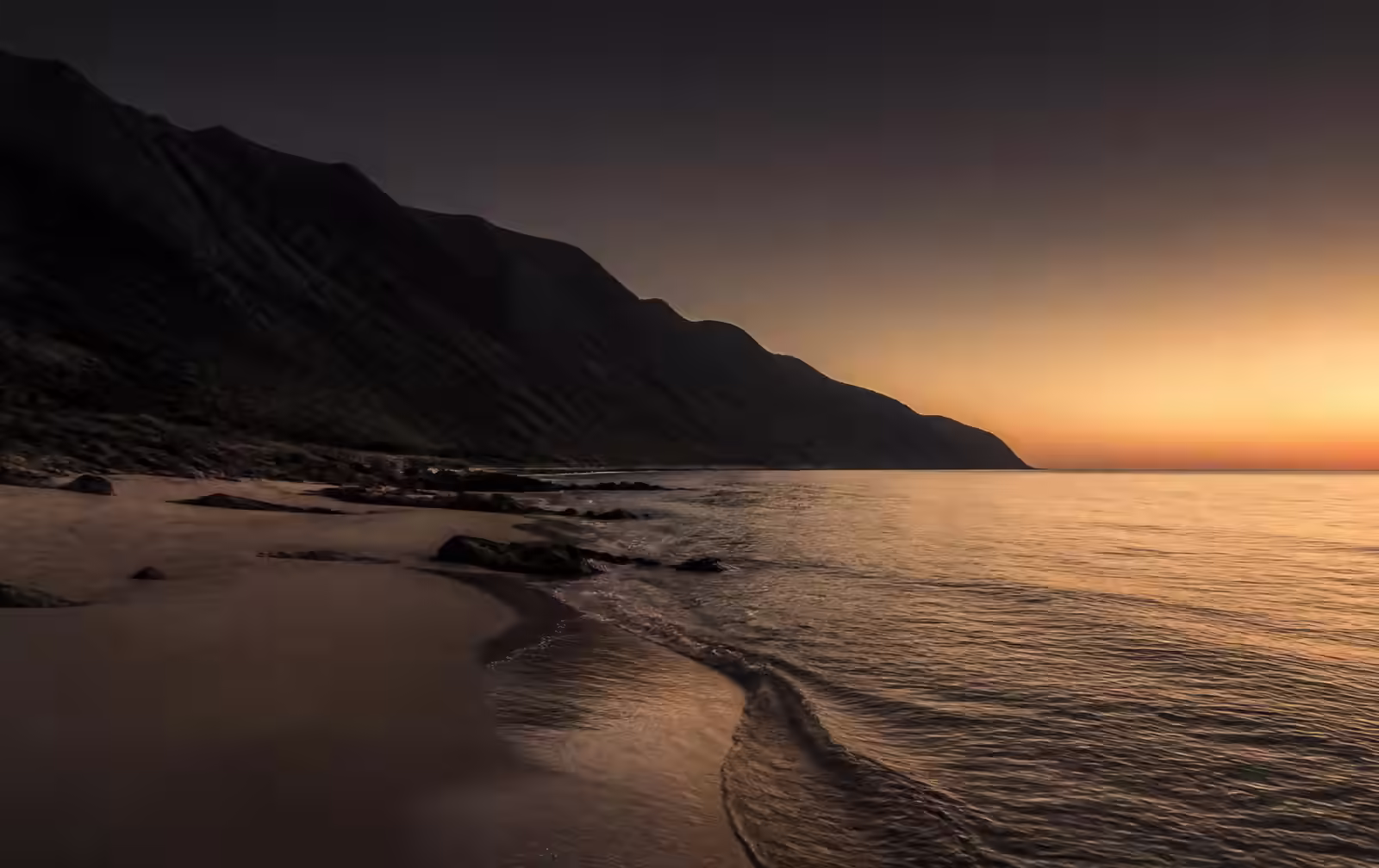 A rocky east-coast shoreline at dusk with dark mountains falling into a calm sea and warm gold horizon light