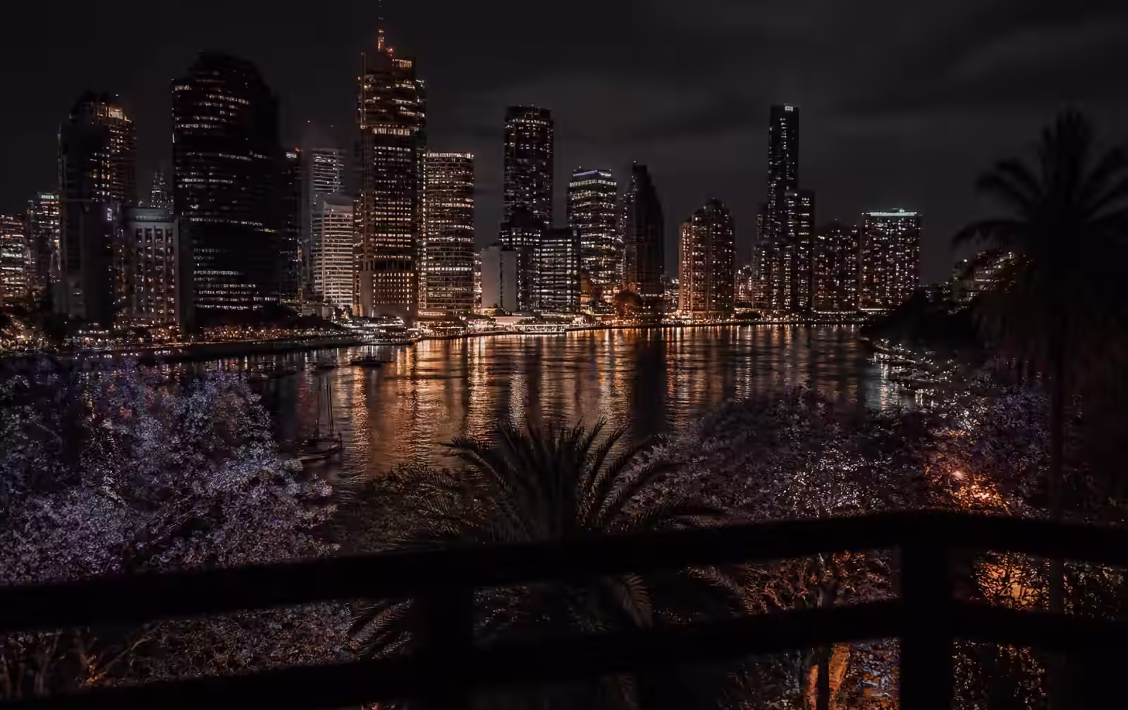 Brisbane's river and glass towers at night with gold reflections on dark water seen past a balcony rail and jacarandas
