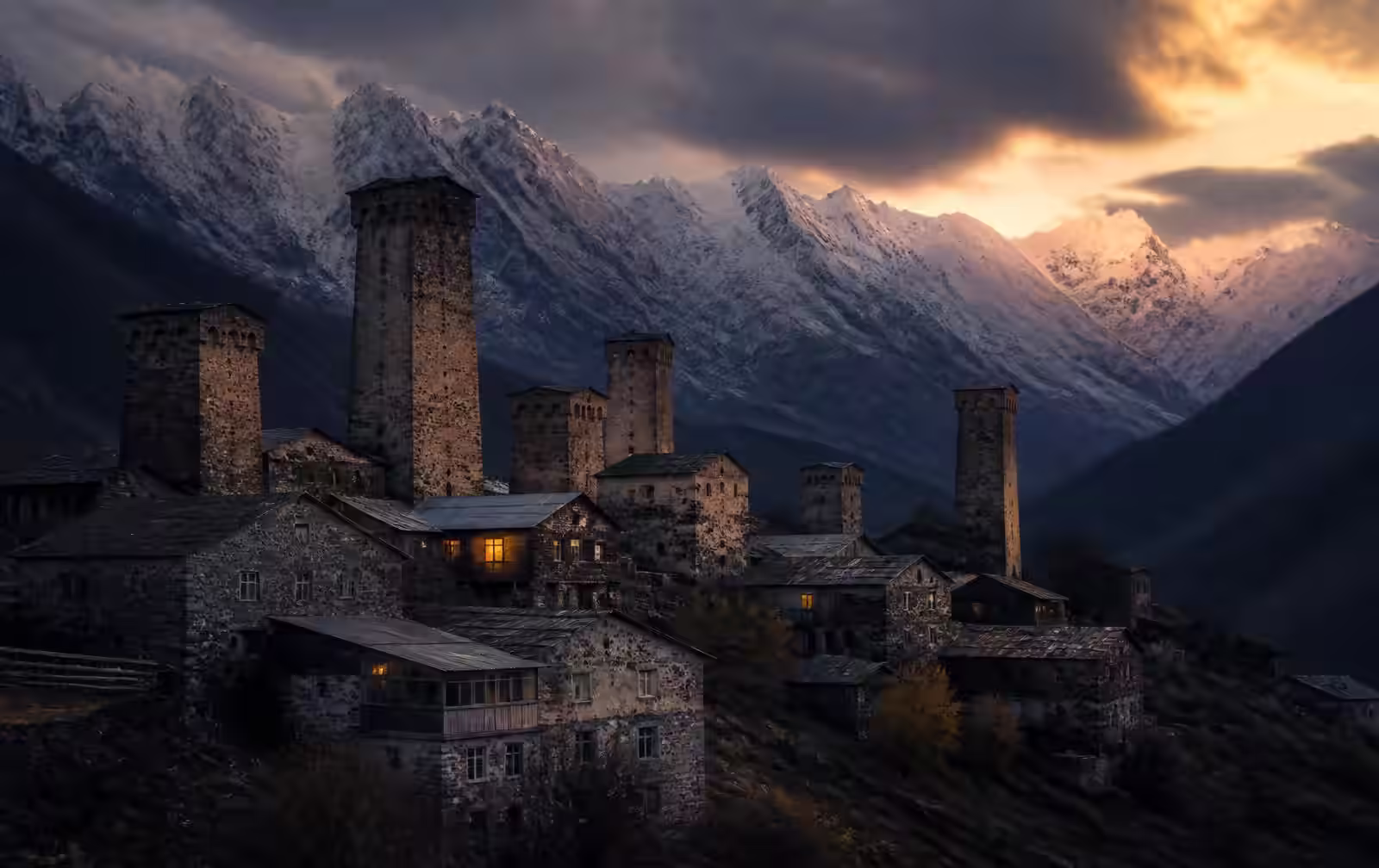 Mestia in Svaneti at dusk, medieval stone watchtowers of a mountain village under snow peaks with a few warm windows