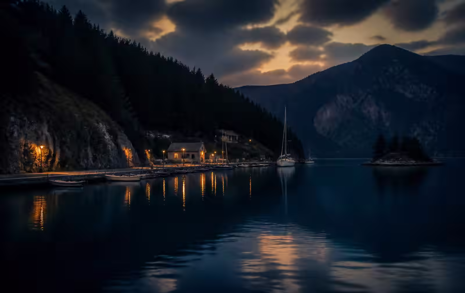 A deep blue harbour at dusk surrounded by dark pine hills with a few warm lights along the quay