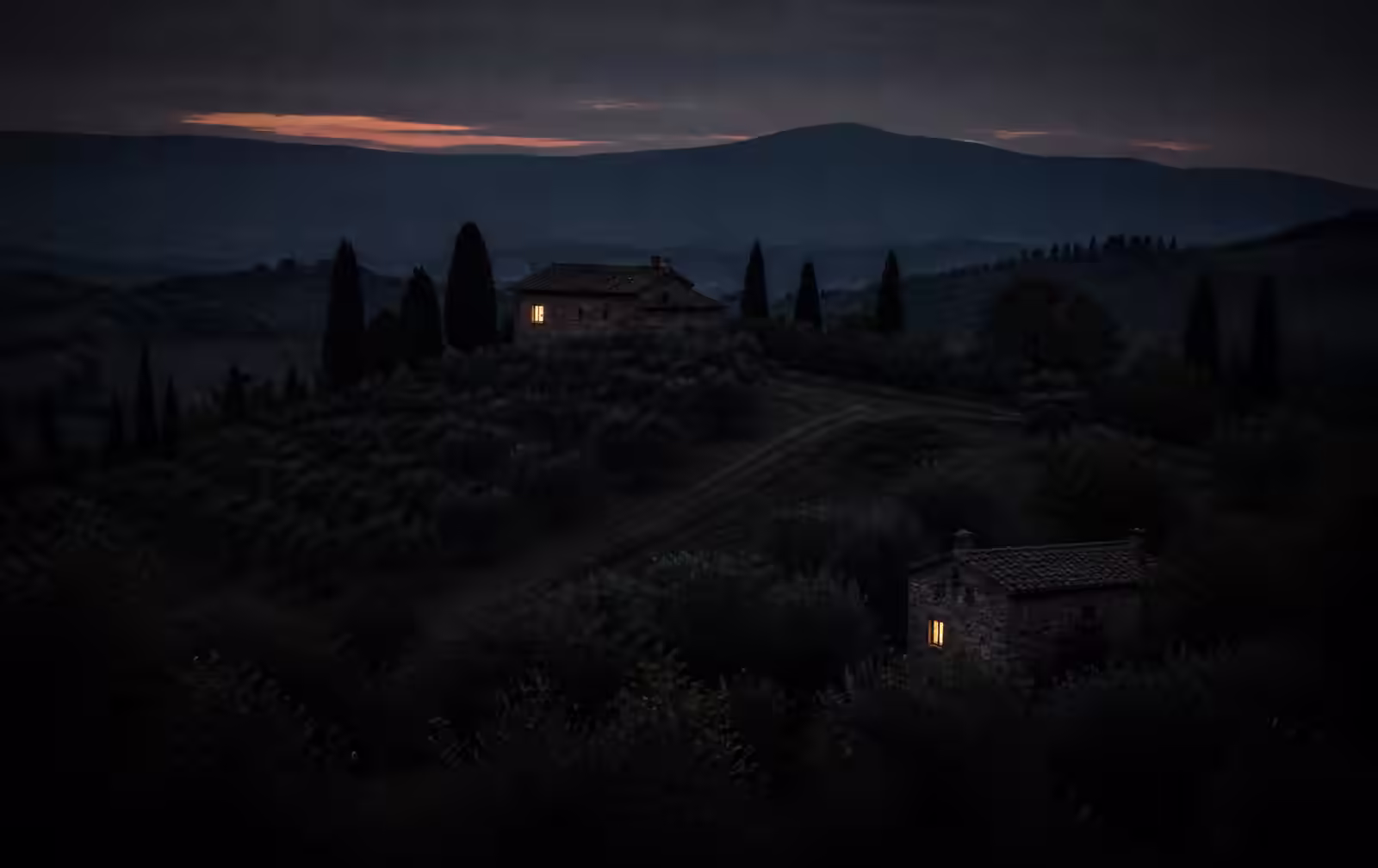 Quiet hills at dusk with two stone houses, a single lit window, cypress trees and layered ridges fading into blue
