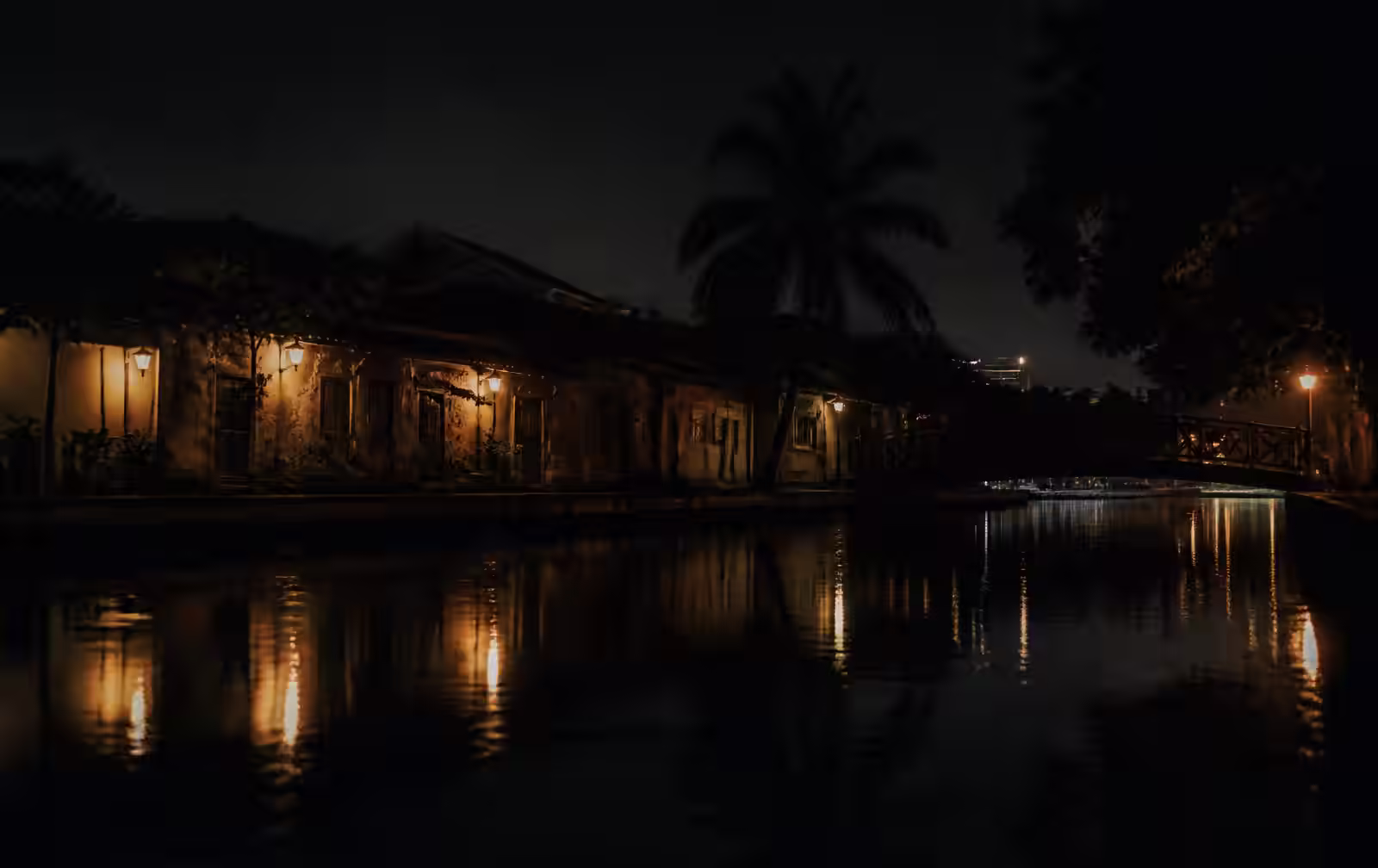A dark riverside at night with warm lamplight reflected on still water, low old buildings and a footbridge in shadow
