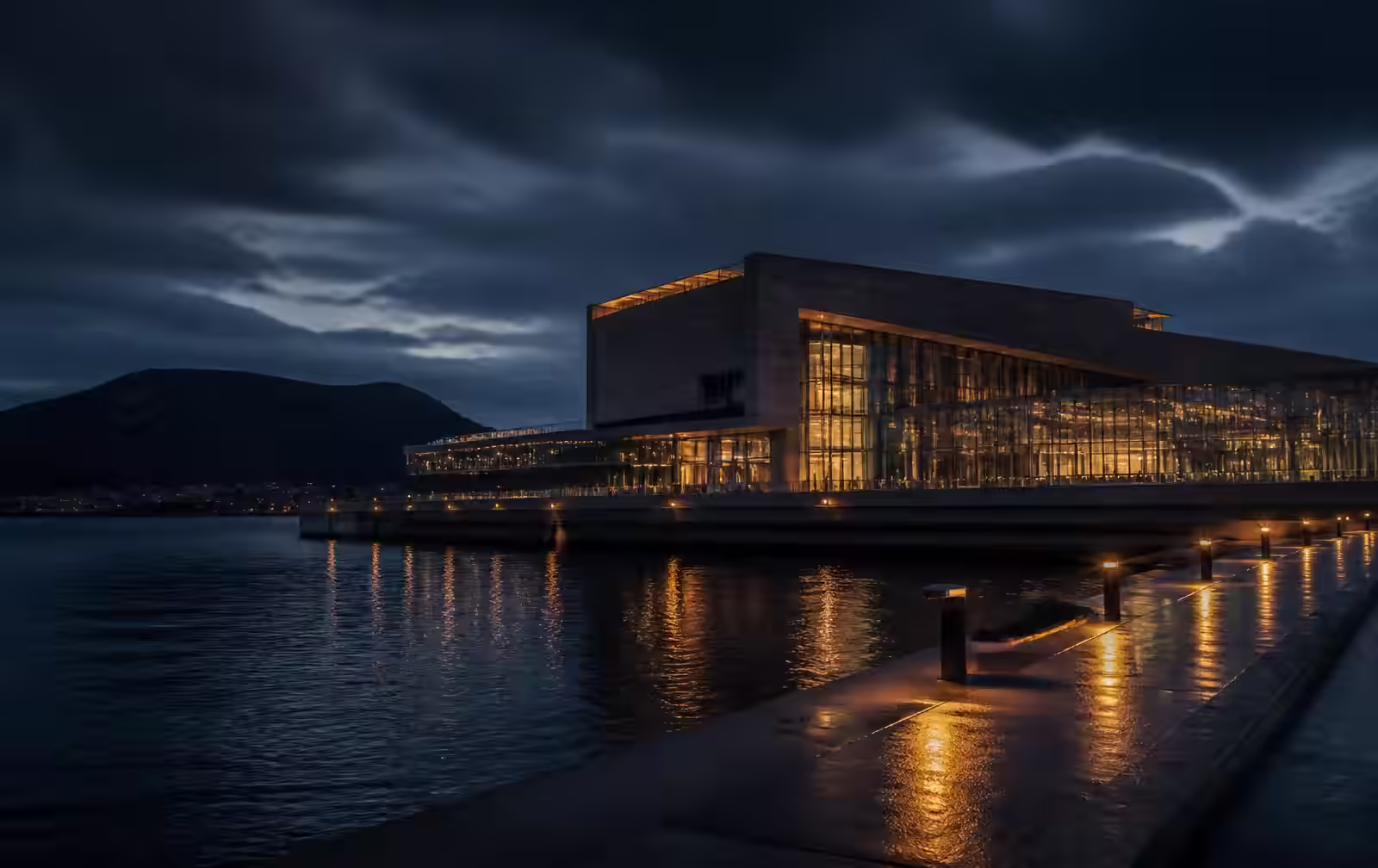 Northern Danish waterfront at night with a modern concrete and glass cultural building lit warmly above dark fjord water and an empty quayside