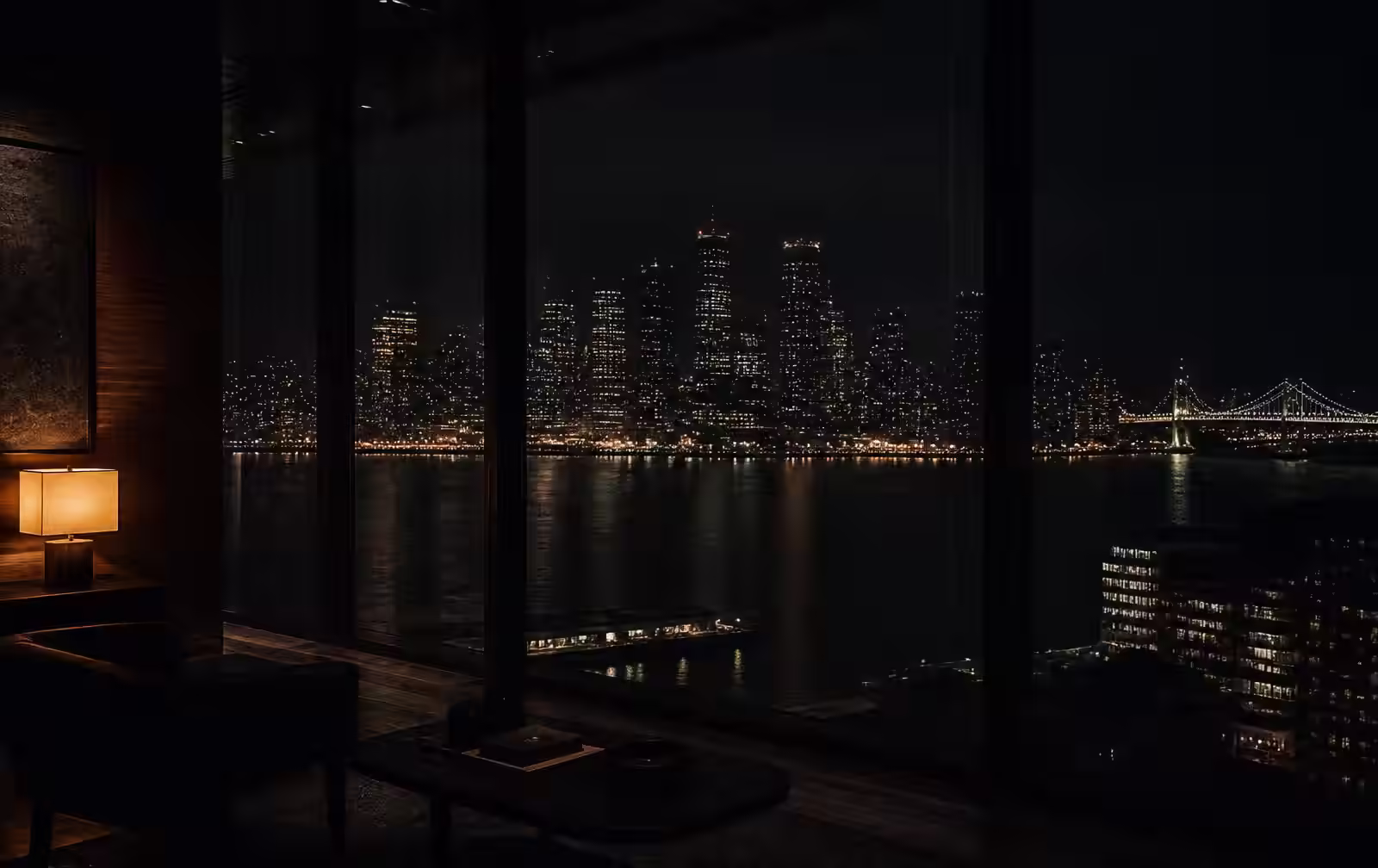A darkened high-rise room with a warm lamp and dark wood, looking through glass at a bay skyline reflected on black water at night