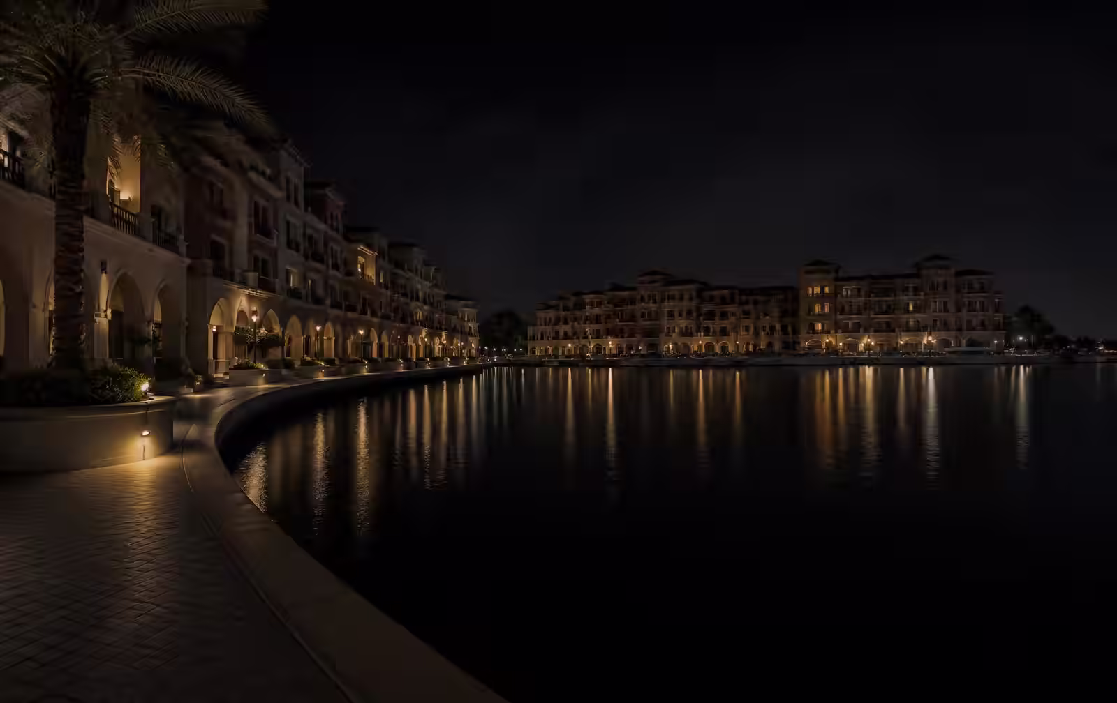 A quiet marina promenade at night, low pastel waterfront buildings darkened, warm lamplight reflected in black water
