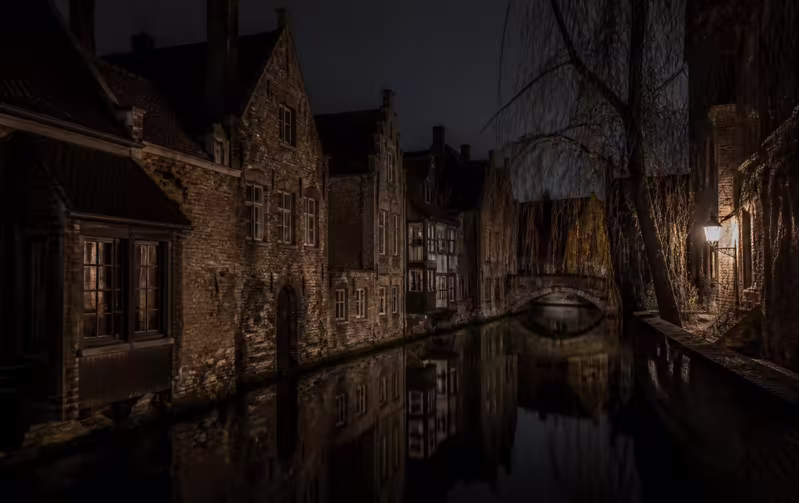 Narrow Bruges canal at night with medieval brick houses, an arched bridge and a single lantern reflected in still water