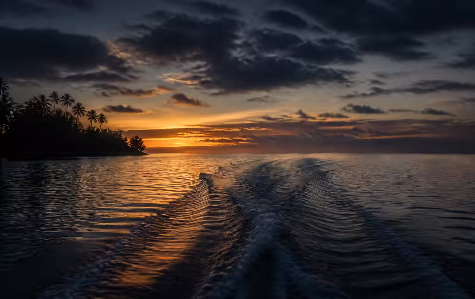A boat wake fading across a calm lagoon at sunset with a palm island silhouetted against gold light