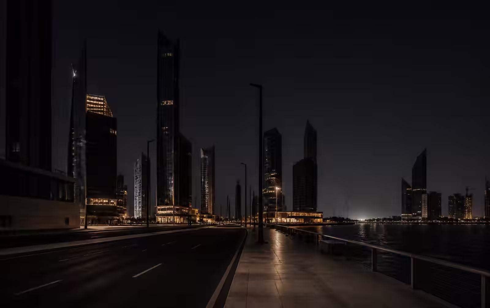 A wide empty waterfront boulevard at night in a very new city, dark high-rise silhouettes with a few warm lit floors beside black harbour water