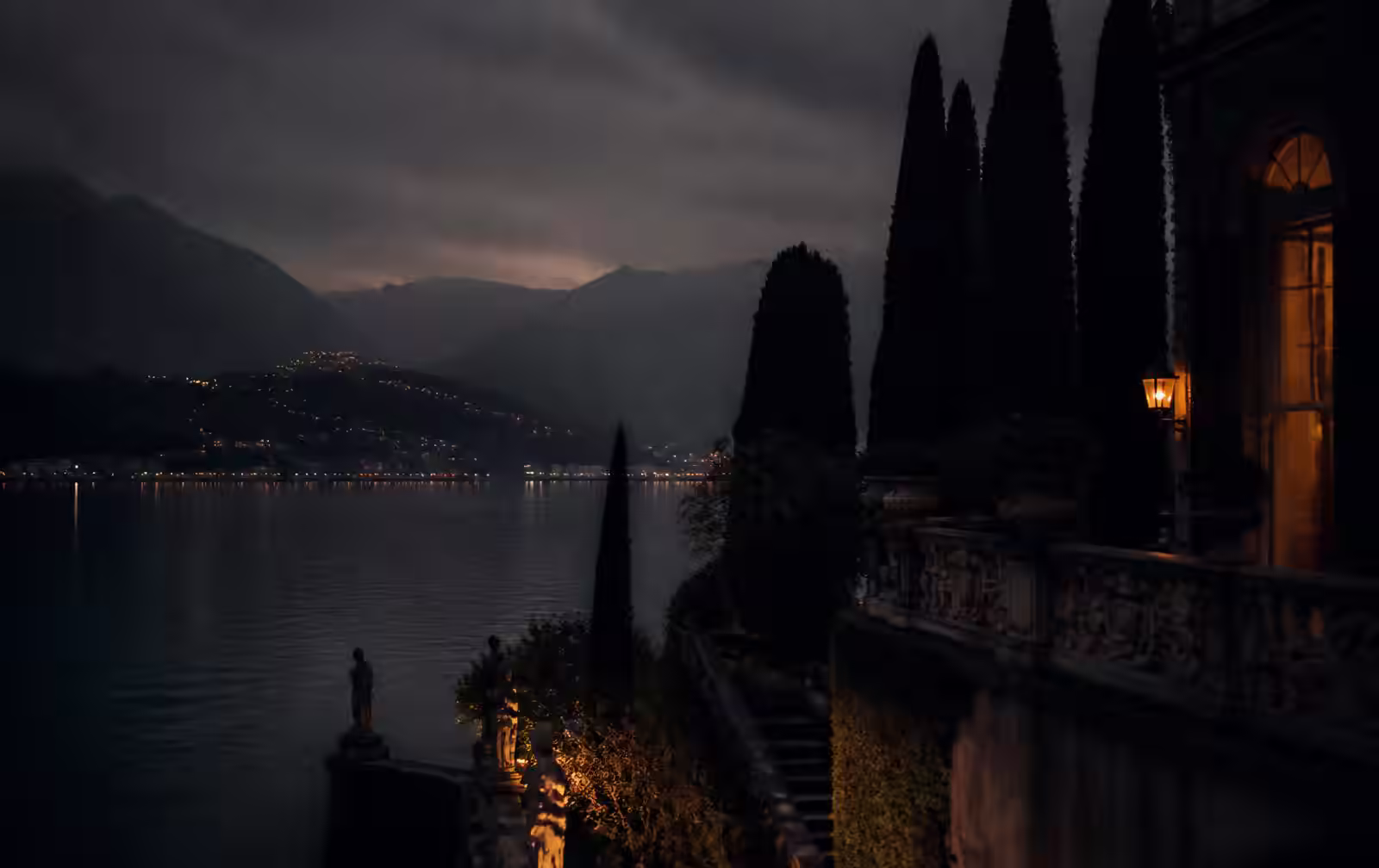 A cypress-lined villa terrace above Lake Como at dusk with lamplight and mist over the dark water