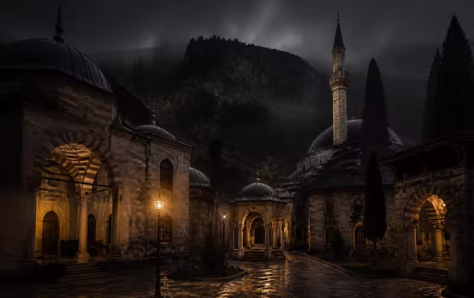 Historic Ottoman-era stone courtyard at night in soft mist with warm lamplight and a forested mountain behind