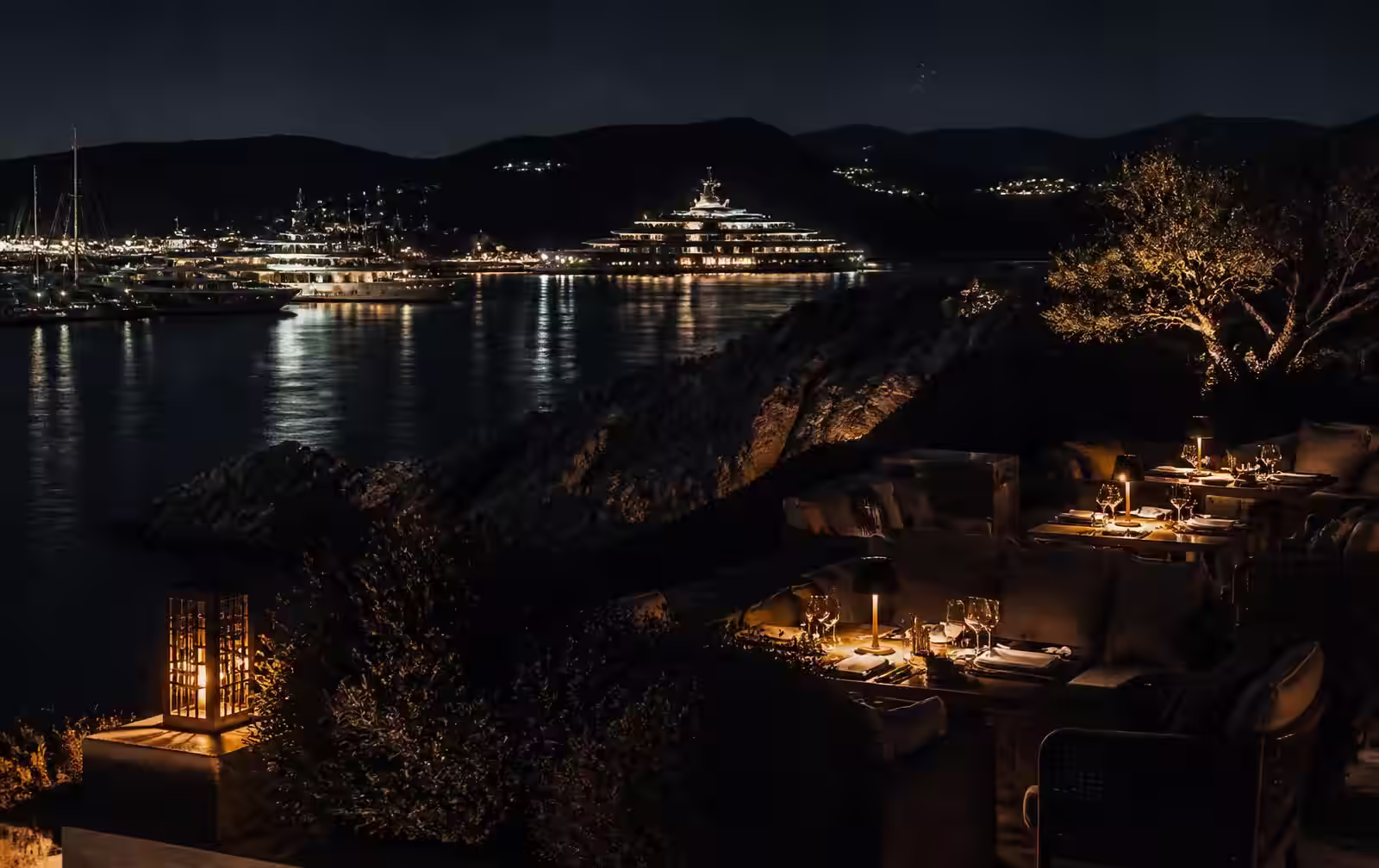 A lantern-lit terrace above a dark Sardinian bay with yachts illuminated in the marina beyond
