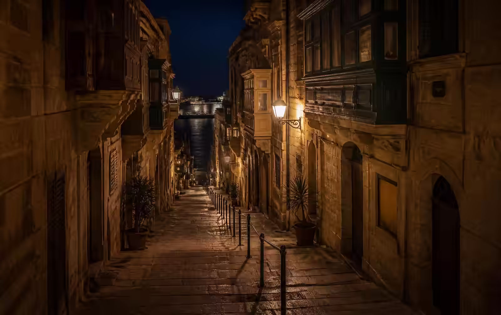 A narrow limestone street in Valletta at night with enclosed wooden balconies, warm lamplight and dark harbour water below