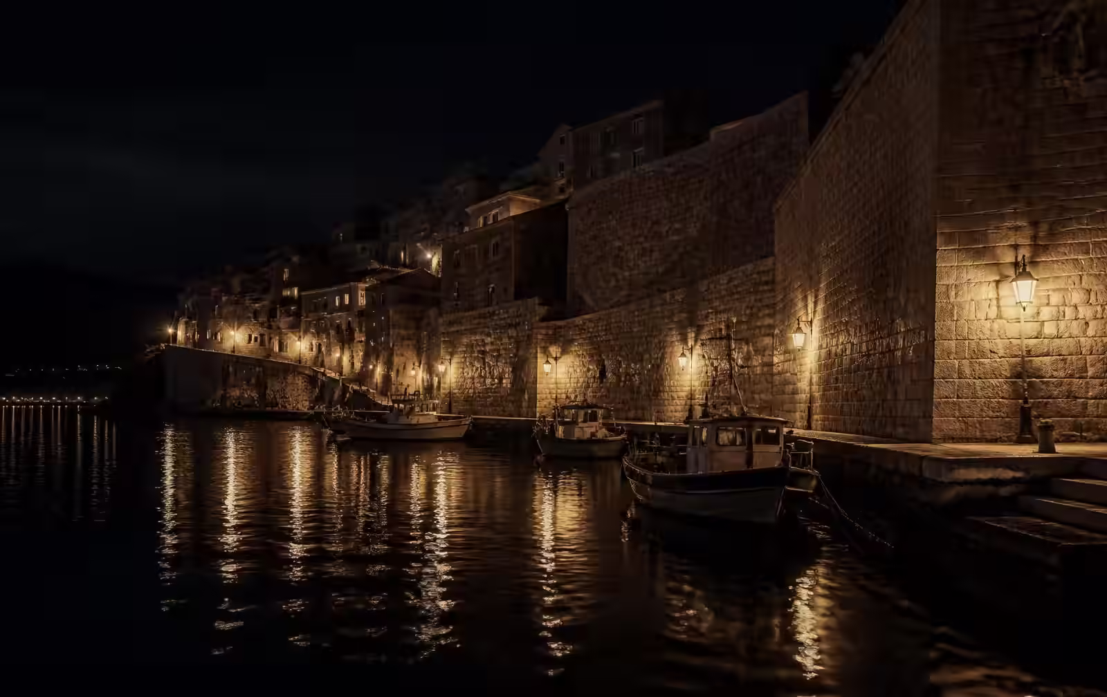 Old stone-walled harbour town at night with lamplit sea walls, moored fishing boats and dark water
