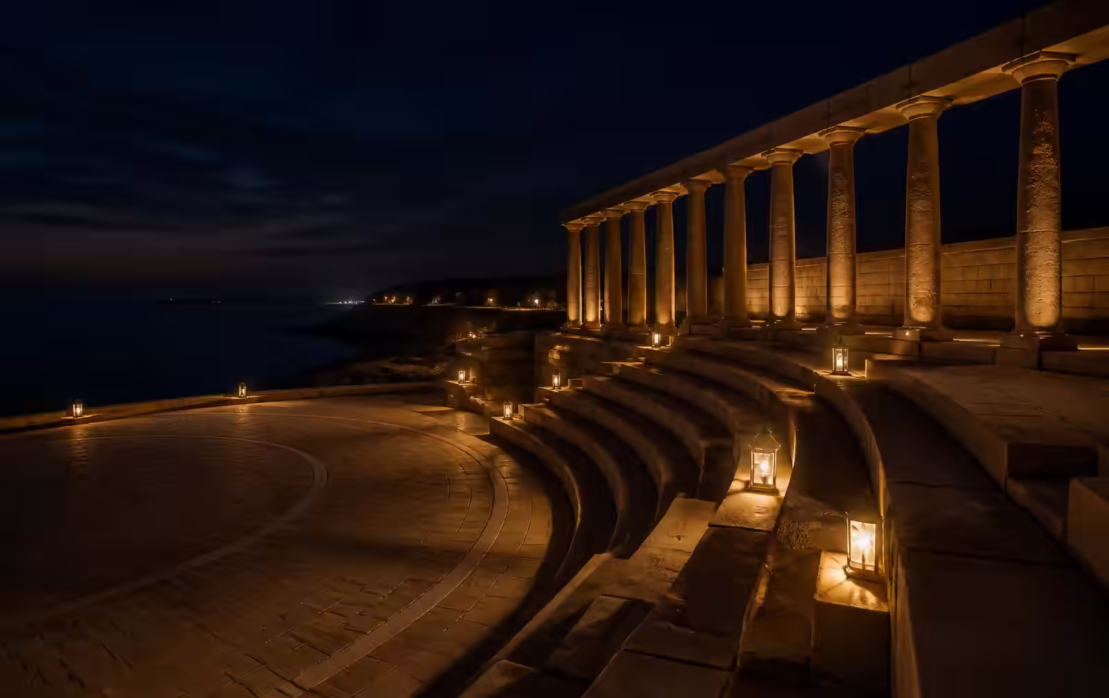 A quiet stone amphitheatre and colonnade beside a dark sea at night, lit by low warm lanterns, entirely empty