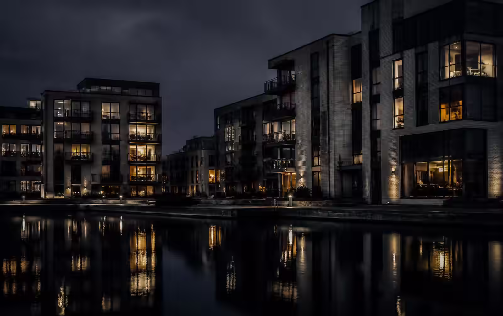 A modern southern Swedish waterfront district at night, contemporary buildings with warm lit windows reflected in a still dark canal basin
