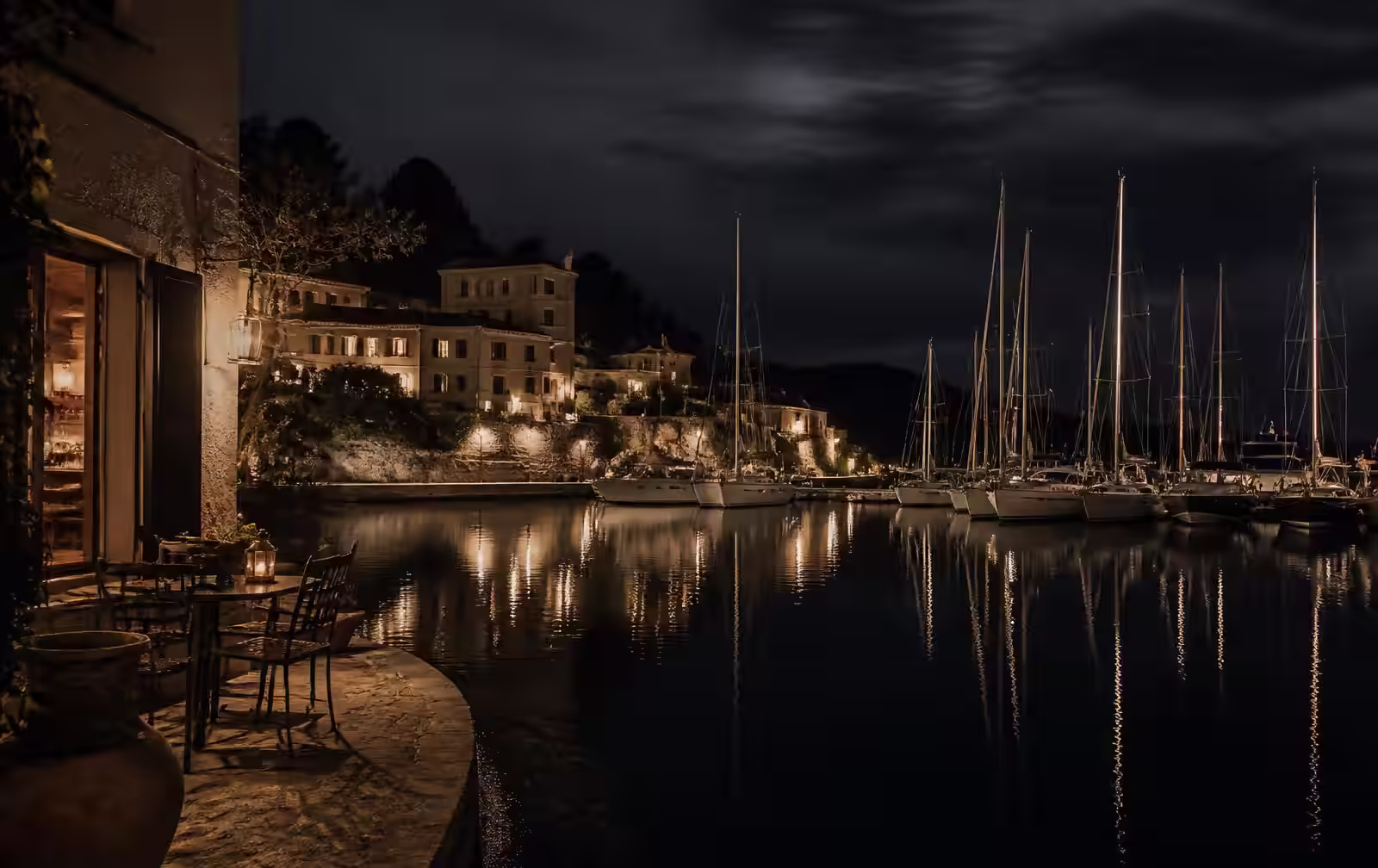 A quiet Mediterranean marina at night with mast lights reflected on black water and a warm coastal interior nearby