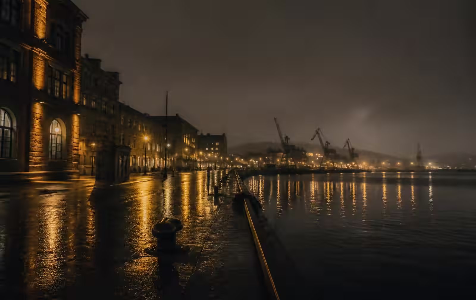 A rain-wet Gothenburg harbour street at night, warm gold streetlights reflected in dark water with distant harbour cranes in fog