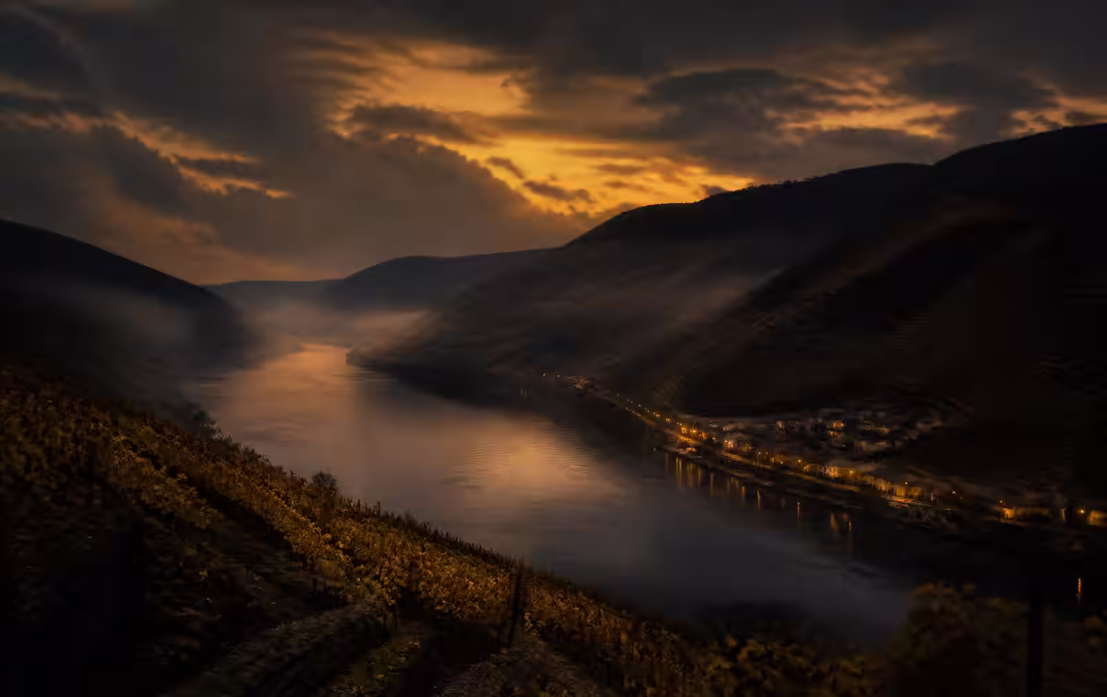 A wide river valley at dusk with terraced vineyards on the slopes and a few warm village lights near the water