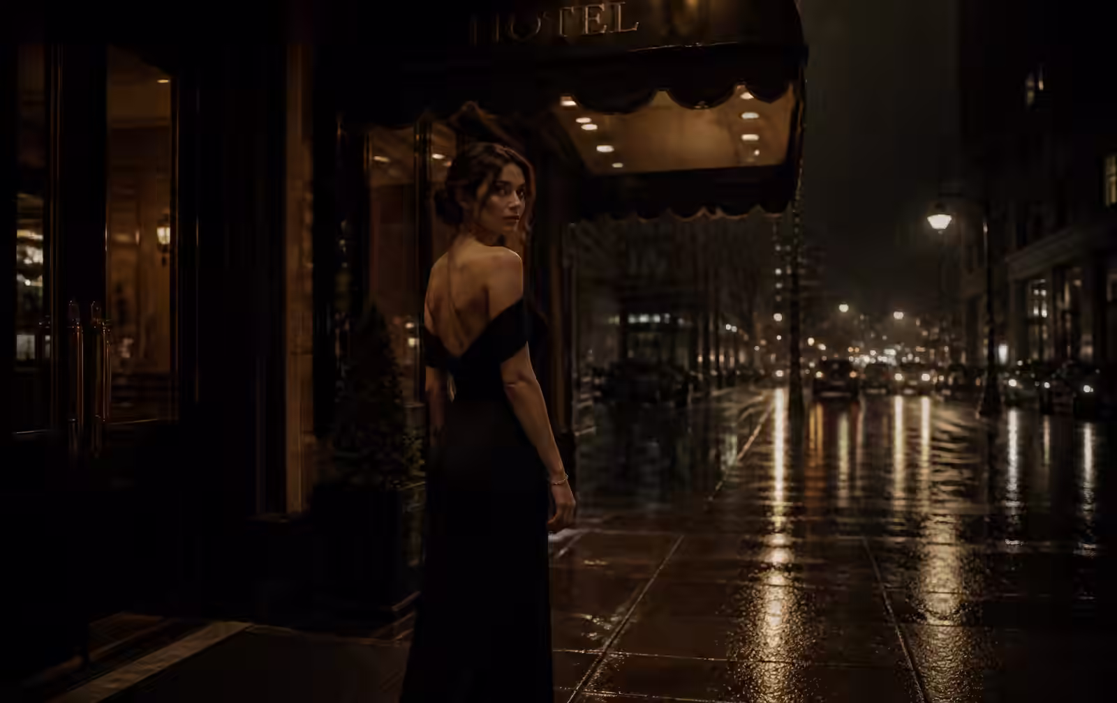 An elegant woman in a black evening dress beneath a hotel awning on a rainy city night