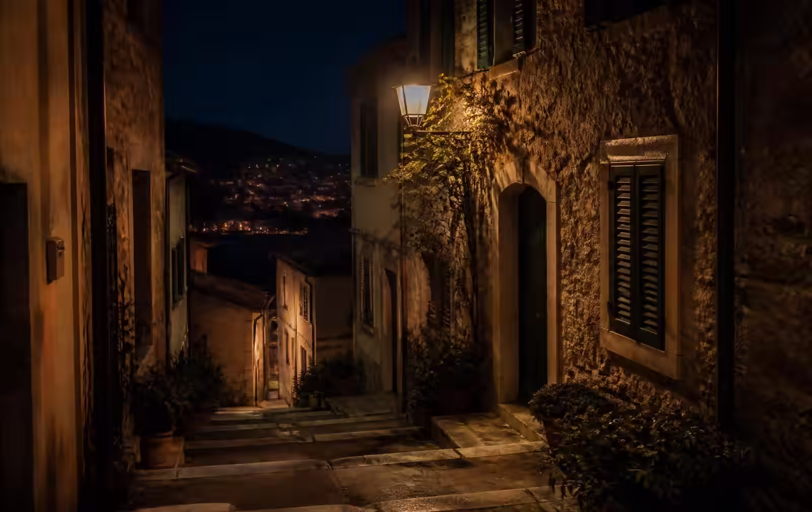 Narrow stepped hillside street at night with pale stone walls, shuttered windows and a single warm lamp