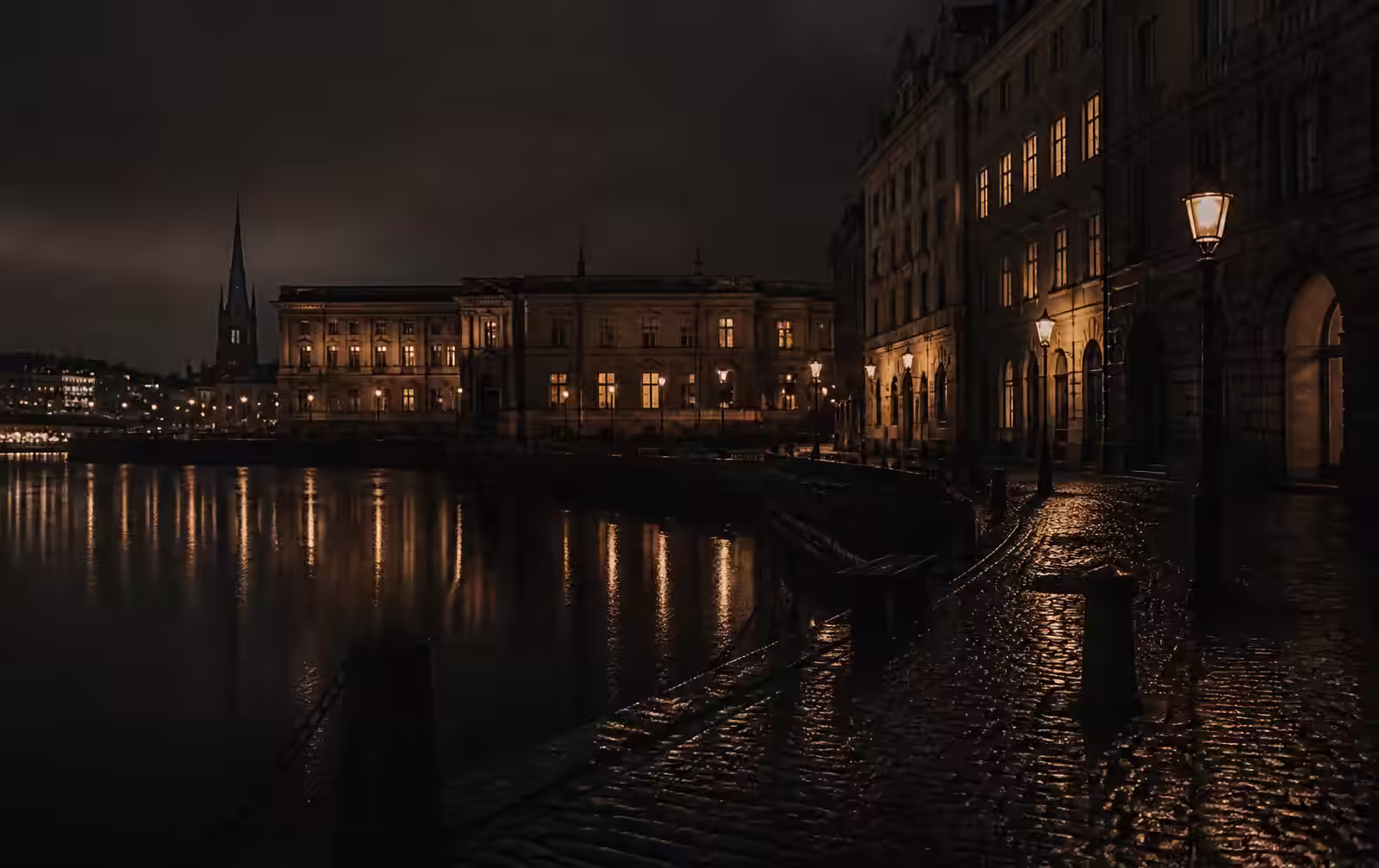 A Stockholm waterfront at night, stone facades with warm amber windows above still dark water and a wet cobbled quay
