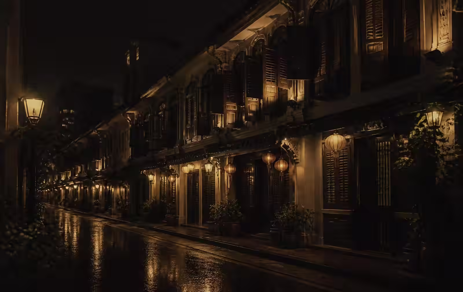 A narrow street of restored shophouses at night with timber shutters, warm lanterns and lamplight reflected on wet pavement